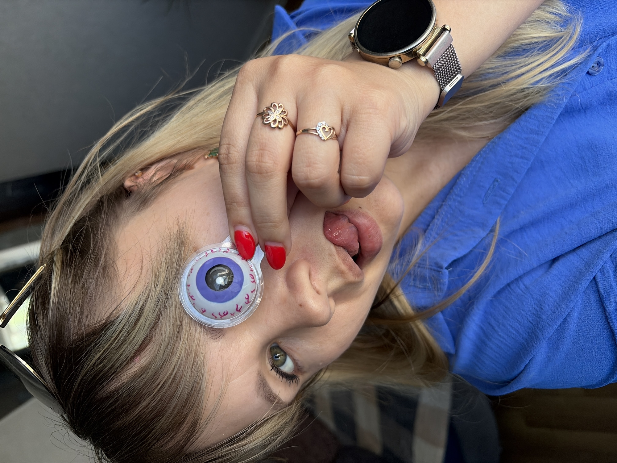 A Caucasian woman with blond hair and green eyes is positioned at an angle in an indoor setting. She is wearing a blue shirt. On her right hand, she has two rings: one in the shape of a flower and another in the shape of a heart. She is also wearing a gold smartwatch with a metallic mesh band on her wrist. In her left hand, she is holding a toy eyeball with purple and white coloring and a black pupil. Her fingernails are painted red. Her tongue is protruding from her mouth. The city location is Chaikovsky, Russia.