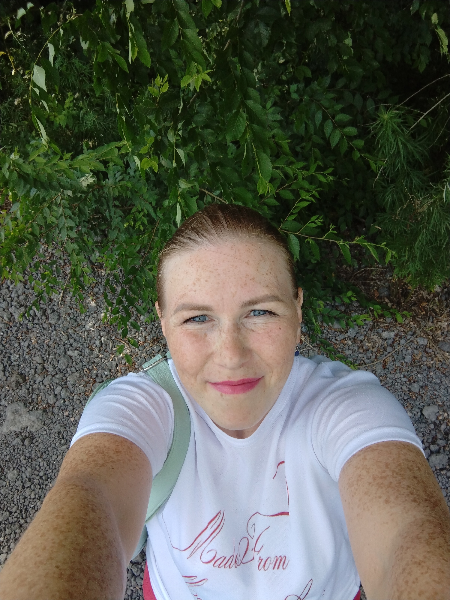 A high-angle, over-the-shoulder selfie shows a Caucasian woman with red hair pulled back, blue eyes, and freckles on her face and arms. She is smiling, with bright pink lipstick. She wears a white t-shirt with a red script graphic and a light green strap is visible over her left shoulder. The background consists of green foliage and gray gravel. The scene appears to be outdoors, possibly in a park or natural area in Komsomolsk-on-Amur, Russia, based on the prompt's implied location.