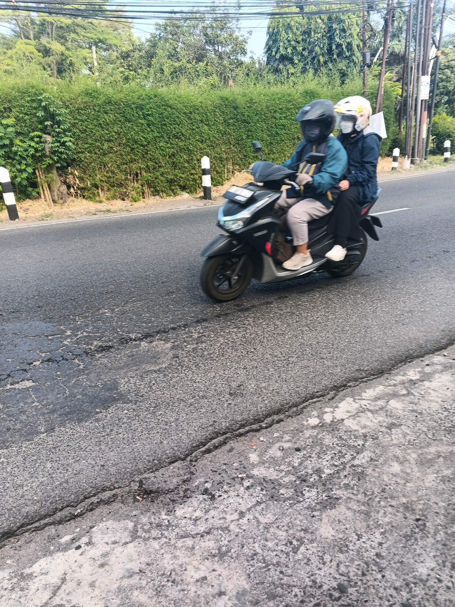 In Ungaran, Indonesia, two individuals are riding a scooter on a paved road. The person in front, wearing a black helmet and a teal jacket, is driving. The passenger behind them wears a white helmet and a dark blue jacket. Both are wearing masks. The road surface shows signs of wear and tear, with patches of cracked asphalt. To the left of the road, a dense green hedge lines the side, with black and white striped posts placed intermittently along the edge. In the background, trees and power lines are visible against a bright sky.