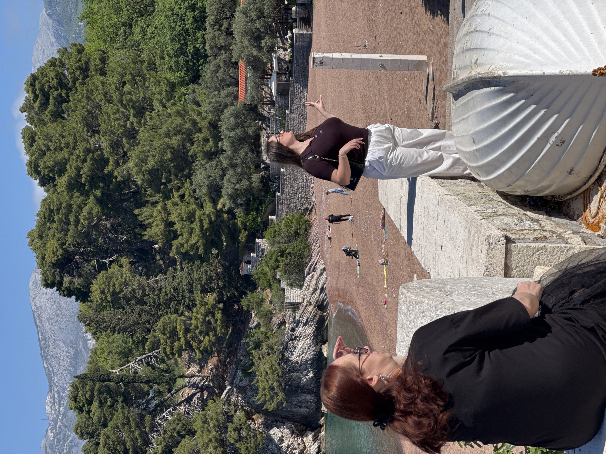 Two women are positioned on a concrete embankment in Sveti Stefan, Montenegro, overlooking a beach. One woman, with brown hair, stands wearing a black shirt and white pants, with a black purse slung over her shoulder. She is pointing towards the beach. The other woman, with red hair, is lying down, wearing a black dress. A white boat is visible on the embankment near the standing woman. The beach below shows several people sunbathing. Beyond the beach is rocky terrain with trees, and a large mountain is visible in the background. The sky is blue.