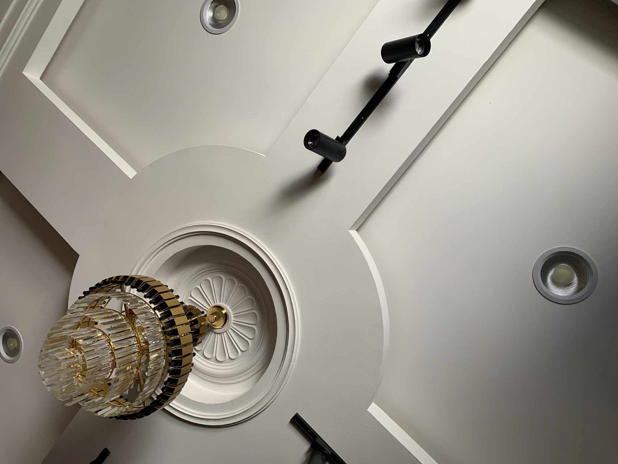 The image shows a close-up, overhead view of a white ceiling with various light fixtures. A crystal and gold chandelier hangs from the center, mounted on a decorative circular molding. To the right, a black track light system with multiple adjustable spotlights is installed. Two recessed circular LED lights are also visible in the ceiling, one in the upper left and another in the lower right. The ceiling has recessed paneling with clean white molding. There is no indication of the city of Gura Top, Nigeria, or any specific location. The scene is static, with no people or actions depicted.