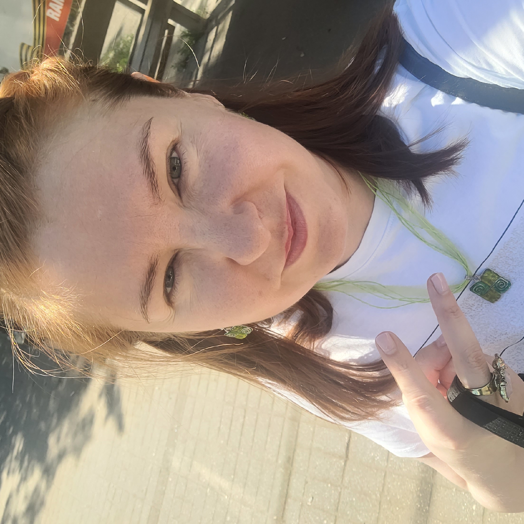 A fair-skinned woman with reddish-brown hair, parted in the middle and pulled back, faces the camera for a selfie in an outdoor, sunlit setting. She has dark eyes and a faint smile, with her lips showing a light pink tint. She wears a white short-sleeved top. On her left ear, a green, spiral-designed dangle earring is visible, matched by another on the right. Around her neck is a light green multi-strand necklace with a square-shaped green pendant featuring a silver border and spiral design. Her left hand is raised, with the index and middle fingers forming a V-shape. Her fingernails are painted a light nude color. She wears a black strap (possibly from a bag or phone) around her fingers and multiple rings, including a gold-colored ring with a light-colored stone and another smaller ring. A black strap from a bag is visible over her right shoulder. The background features light-colored paved ground with dappled shadows from unseen foliage on the left. On the right, a blurry structure resembling a wooden fence or railing is visible, along with a red and white sign displaying partial letters "RA" (possibly "RAM"). The scene is brightly lit by natural sunlight, indicating it is daytime in the city of Unknown, Unknown.
