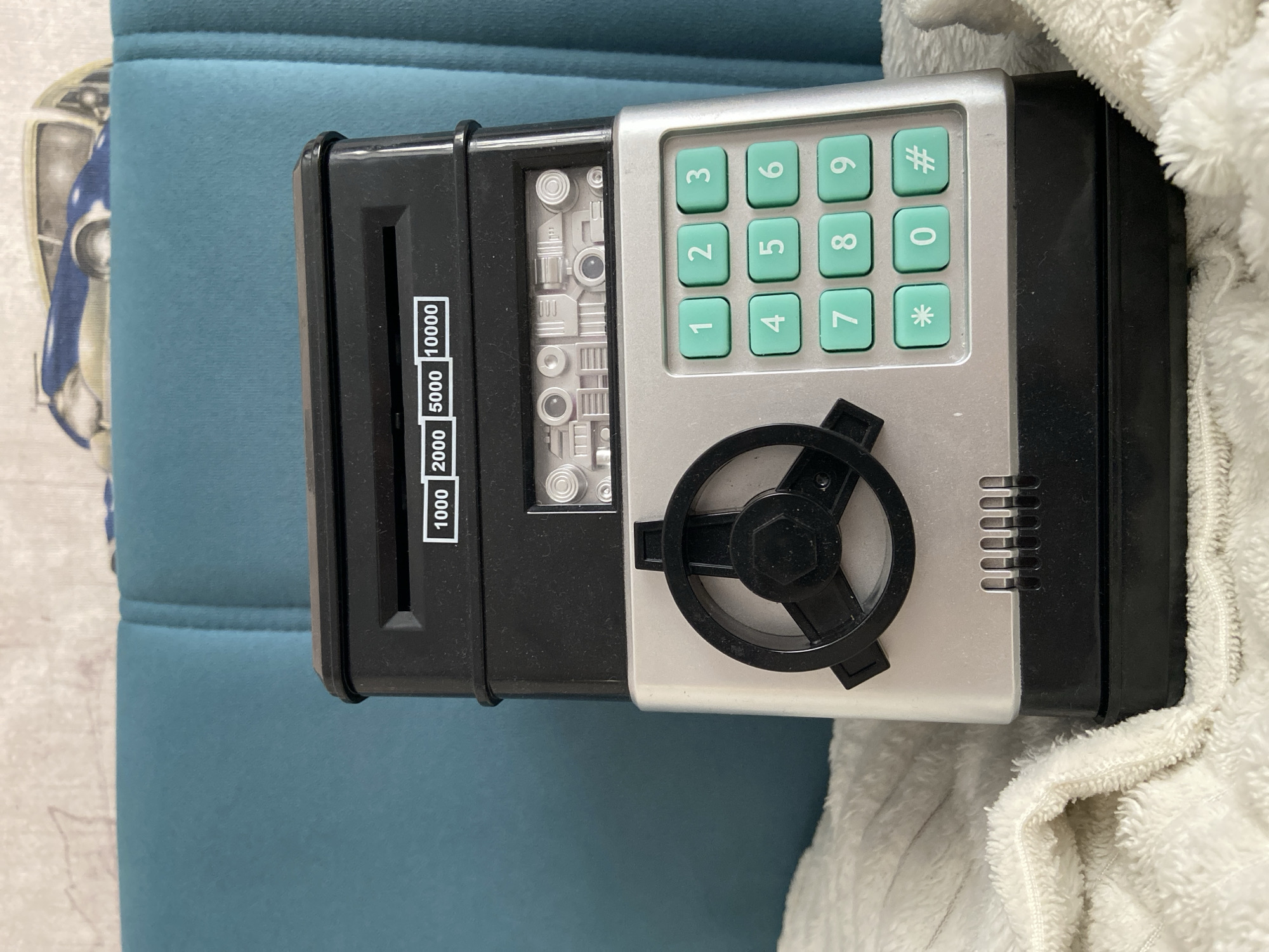 A black and silver electronic piggy bank, designed to resemble a safe, sits on a white textured blanket. The piggy bank has a slot at the top for inserting money, with labels indicating denominations of 1000, 2000, 5000, and 10000. Below this is a panel with a decorative metallic design, resembling mechanical parts. The lower section features a circular dial with a black handle and a keypad with green buttons numbered 0 through 9, along with asterisk and hash symbols. The background consists of a teal-colored upholstered surface, likely furniture, with a patterned fabric featuring a vintage car visible at the top. The scene is indoors, on a bright day, and the location is Balashikha, Russia.