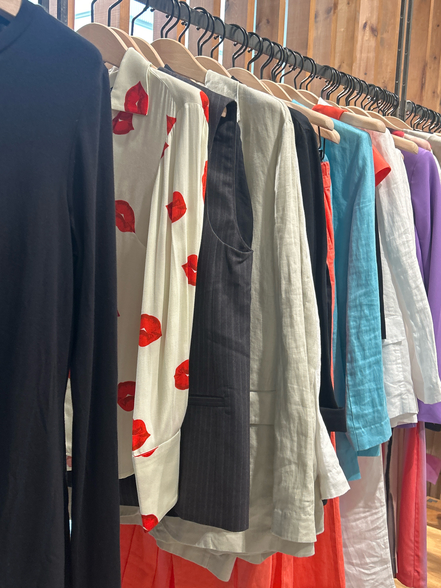 A rack of women's clothing is displayed against a wooden wall. The clothing items, from left to right, include a long-sleeved black garment, a cream-colored shirt with a red lip print pattern, a dark pinstripe vest, a light beige linen jacket, a black top, an orange skirt, a bright blue linen top, a white linen jacket, and a purple jacket. The clothes are hung on wooden hangers. The background consists of vertical wooden planks. The scene appears to be in a retail environment, possibly a boutique or thrift store. The location is stated to be Gamarjveba, Georgia.