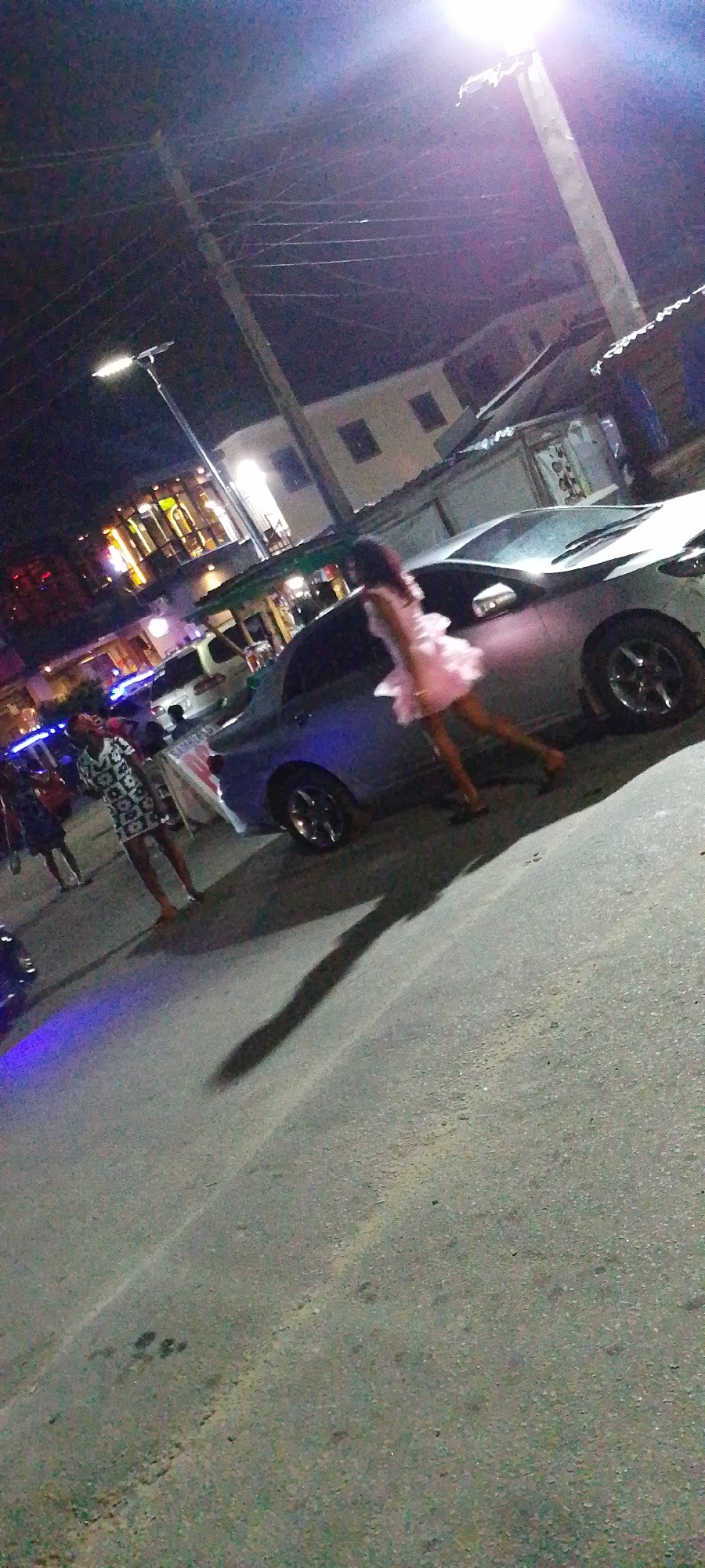 The image shows a street scene at night in Lokoja, Nigeria. A car is parked on the side of the road near a building. Several people stand and walk nearby. Overhead, there are electric power lines. The lighting is provided by street lamps.