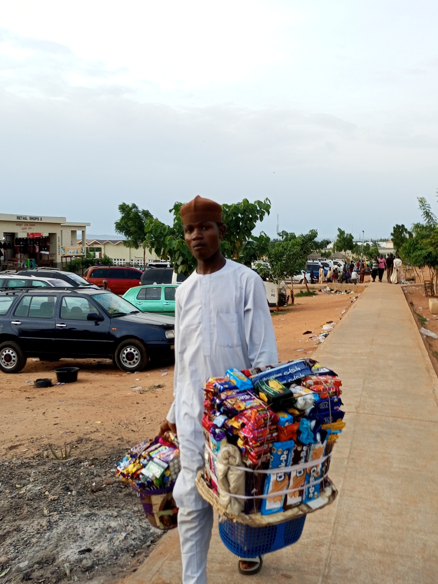 A man wearing a white traditional outfit and a brown cap stands in the foreground of the image, carrying two large baskets filled with various packaged snacks. He is walking on a paved walkway. The baskets are stacked high with colorful wrappers of cookies, biscuits, and other confectioneries. The man is looking directly at the camera. In the background, several cars are parked on a dirt lot, and a building with "RETAIL SHOPS 2" visible on it is in the distance. There are trees and other people in the background. The sky is overcast. The scene appears to be an outdoor market or commercial area in Gombe, Nigeria.