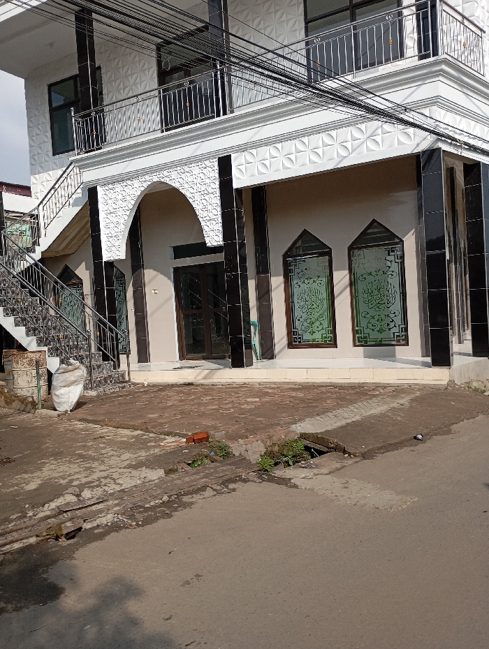 An exterior daytime photograph captures a multi-story building, likely a mosque or Islamic center, situated in Situwangi, Indonesia. The structure features a white facade with recurring geometric patterns on its upper sections and black polished accents, including vertical pillars and window frames. The ground level includes a central arched entryway adorned with white intricate patterned relief, containing dark double doors. To the right, two vertically oriented, arch-topped windows display green-tinted glass etched with white Arabic script. An external staircase with metal railings ascends on the left, leading to a second-floor balcony also equipped with metal railings. Multiple black overhead utility lines are visible traversing the upper portion of the frame. The immediate foreground comprises a section of reddish-brown brick pavement, transitioning to a worn asphalt street with a visible concrete drainage channel. Proximate to the staircase, two rusted metal drums and a large white fabric-like bag are present. A single red brick lies on the pavement near the drainage. Minimal vegetation growth is observed within pavement cracks and the drainage channel. No human subjects are present, and no discernible actions or interactions are depicted within the static scene.