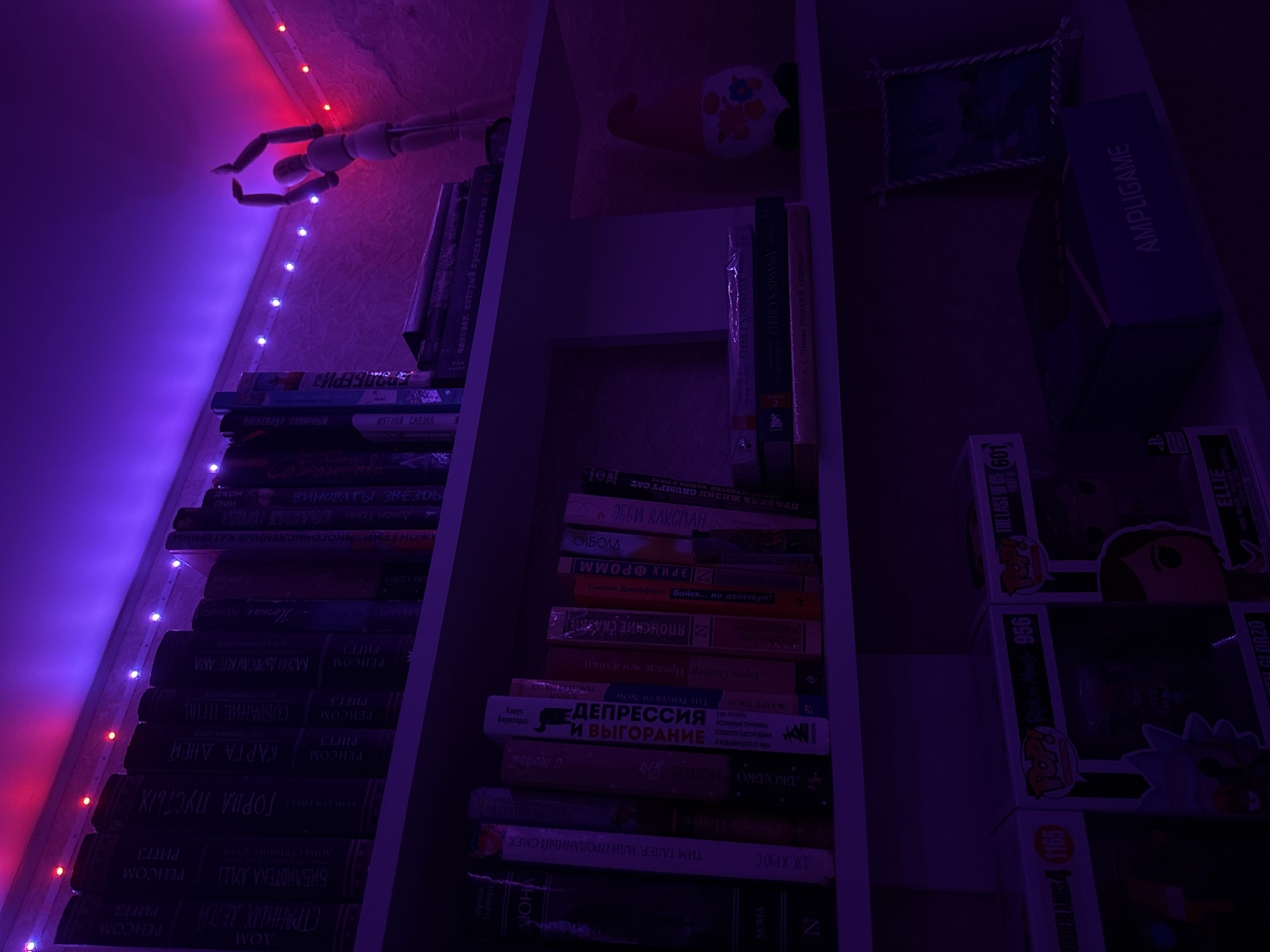 The image shows a bookshelf in what appears to be a dimly lit room, likely in Pudomyagi, Russia. The shelves are filled with books, and decorative items. The room is illuminated by purple and pink LED strip lights. On the top shelf, there is a wooden mannequin in a dancing pose. The middle shelf contains various books, including titles like "Depression and Recovery" and "The Power of Now." On the bottom shelf, there are Funko Pop figures, a framed picture, and a box with "AMPLIGAME" written on it. A gnome figurine is also present on the bottom shelf.
