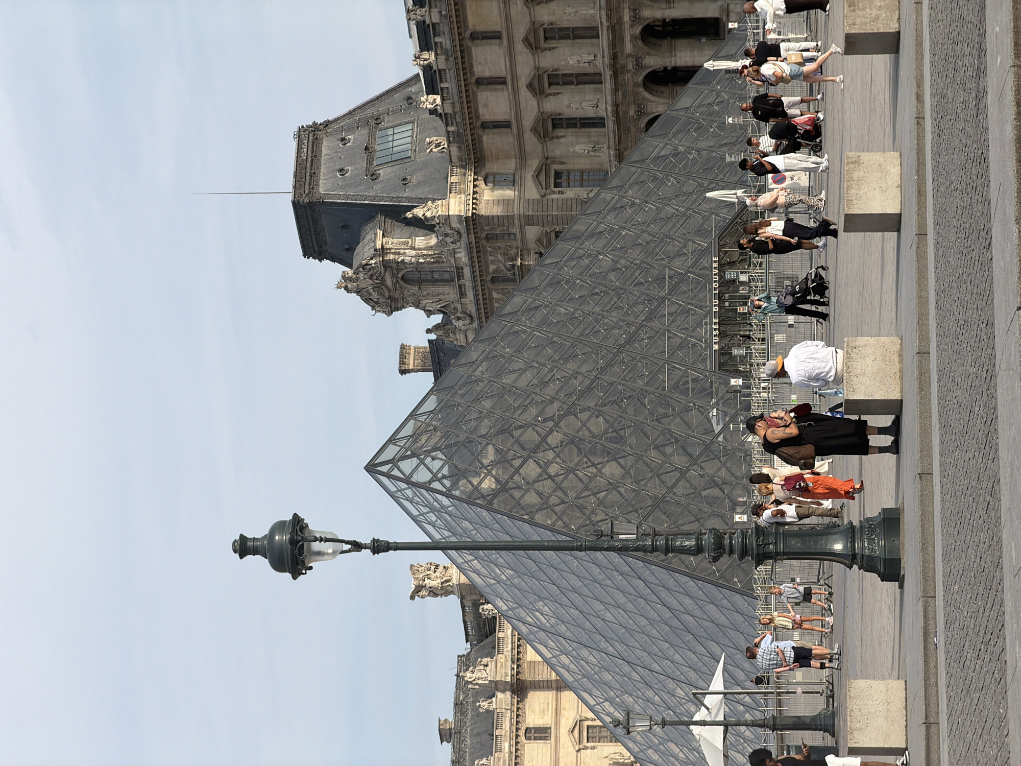 This is a daytime, eye-level, full shot of the Louvre Pyramid in Paris, France. The iconic glass pyramid is the central element of the image, reflecting the sky and surrounding buildings. In the foreground, a detailed black lamppost stands prominently, adding a historical architectural element. Several people are scattered across the plaza in front of the pyramid, some walking, some standing and conversing, and one person sitting on a concrete barrier. The Louvre Museum building is visible behind the pyramid, showcasing its classical architecture. The ground is paved with cobblestones, and large concrete blocks are placed as barriers in the foreground. The sky is a clear, pale blue, indicating fair weather. The overall atmosphere is that of a tourist destination with people enjoying the iconic landmark.