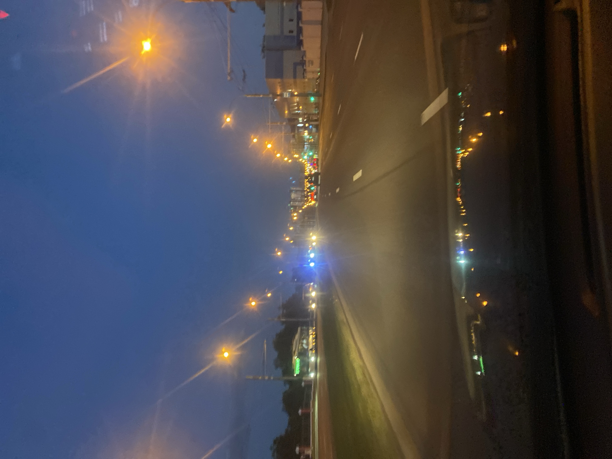This is a photograph taken at dusk from inside a vehicle traveling on a multi-lane road. The sky is a deep blue, transitioning from twilight. Streetlights with orange and yellow hues illuminate the scene, casting long streaks of light due to the movement and low light conditions. On the left side of the road, there is a grassy median and trees. Several vehicles are visible on the road, including cars with red taillights and what appears to be a bus with illuminated green signage. The road surface shows painted white lane markers. Buildings, some with illuminated windows, are present on the right side of the road. The foreground shows the reflection of lights on the car's dashboard and windshield. This appears to be a street scene in Minsk, Belarus, likely along a major thoroughfare during the evening commute or after dark.
