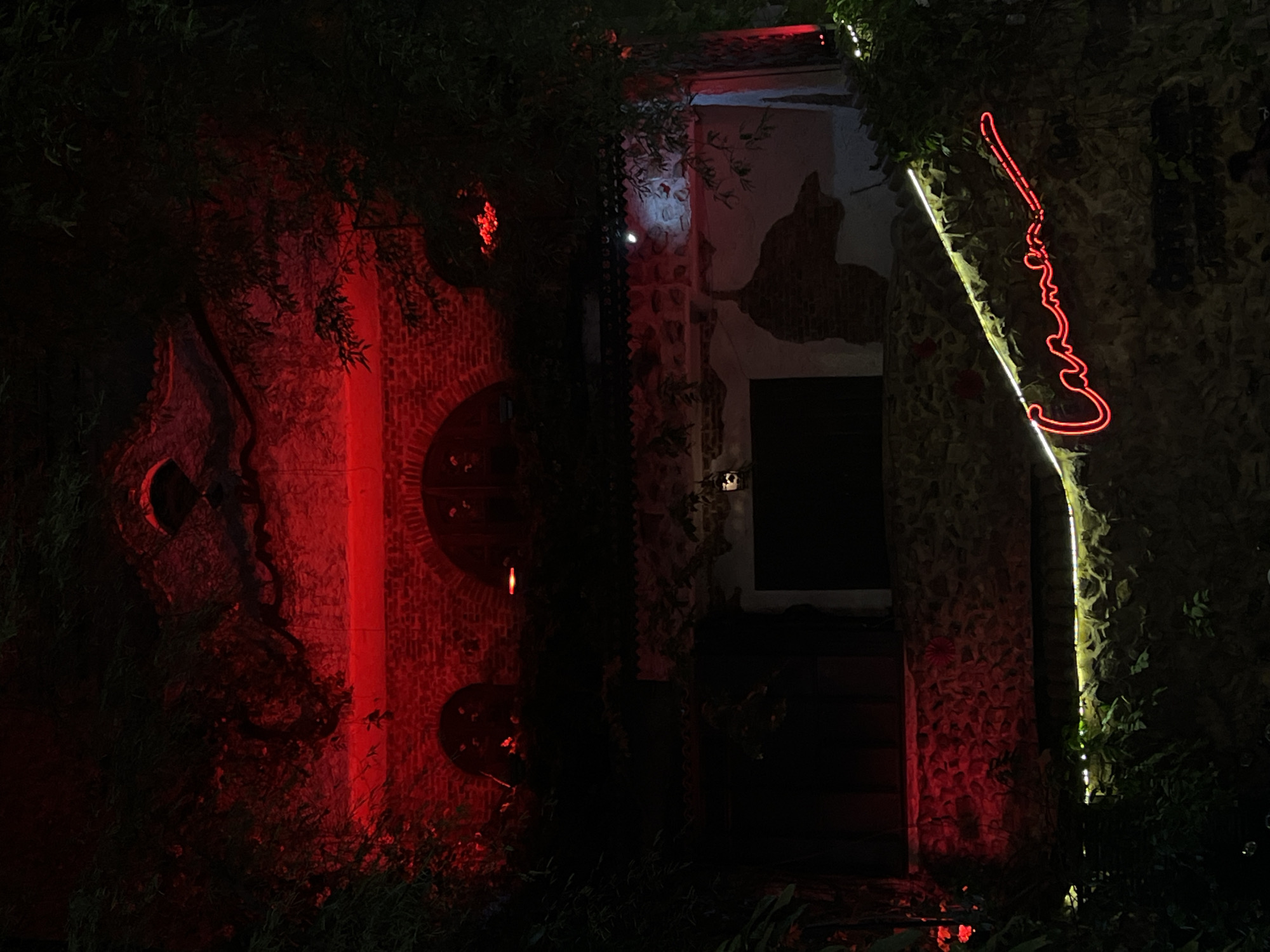 The image shows a dimly lit exterior of a building, likely at night, in Thủ Đức, Vietnam. The building is constructed with stone and brick, and is partially covered in foliage. Red lights illuminate the upper facade, creating a dramatic effect. On the lower part of the building, a neon sign in cursive script glows red, spelling out "Carmen". The sign is positioned diagonally across a stone wall. White LED strip lights run parallel to the neon sign. The overall atmosphere is dark and moody, with the vibrant red lights contrasting with the shadowed surroundings and greenery.