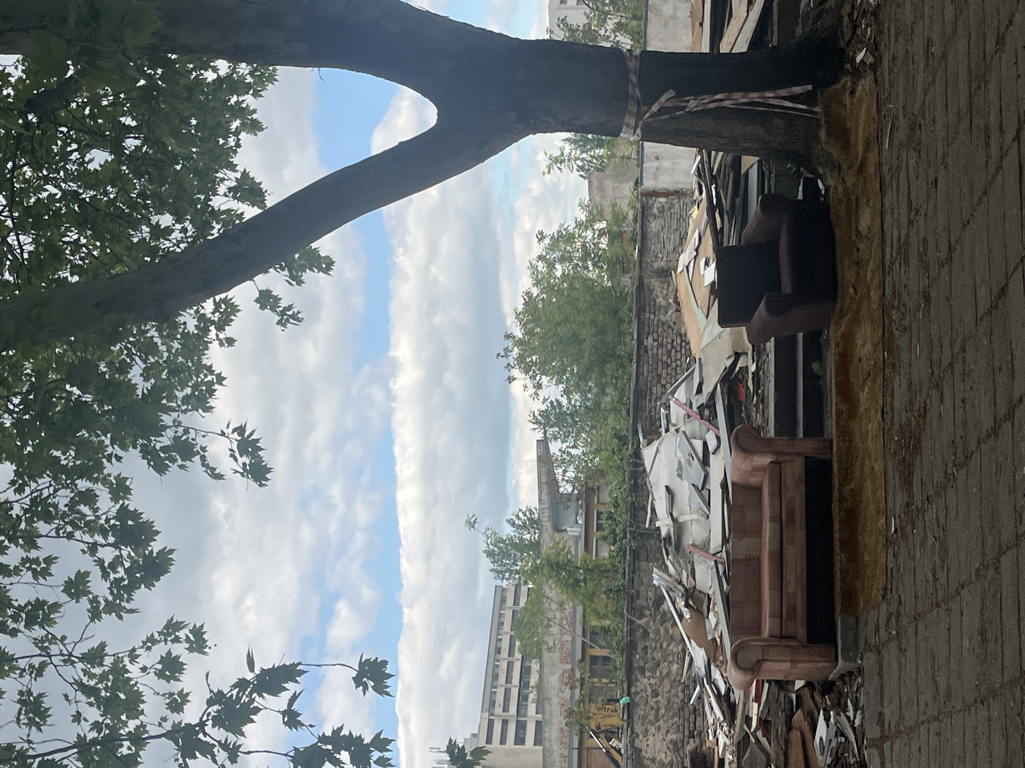 The image shows a partially obstructed view in Plovdiv, Bulgaria. A tree with green leaves occupies the left portion of the frame, partially obscuring the sky with scattered clouds. In the foreground on the right side is a paved sidewalk. Beyond the tree and sidewalk, a pile of debris is visible, including furniture such as sofas and various construction materials. Behind the debris, there are buildings of varying heights, suggesting an urban environment. A small, yellow construction vehicle is visible amidst the debris near a partially ruined wall.