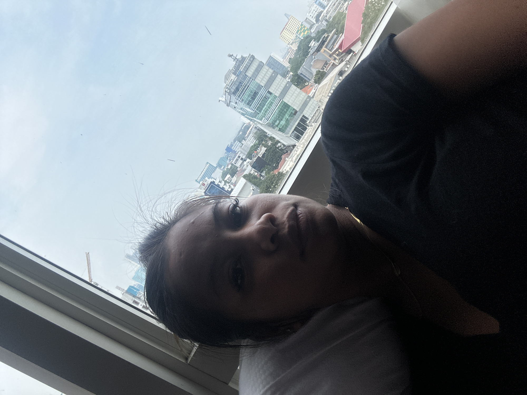 The image depicts a woman indoors, likely in a high-rise building, looking out of a window. She has dark hair and skin, and is wearing a black shirt. The view from the window includes several tall buildings, suggesting an urban setting. Construction is visible in the distance. Based on the architecture and available information, the location is likely Central Jakarta, Indonesia.