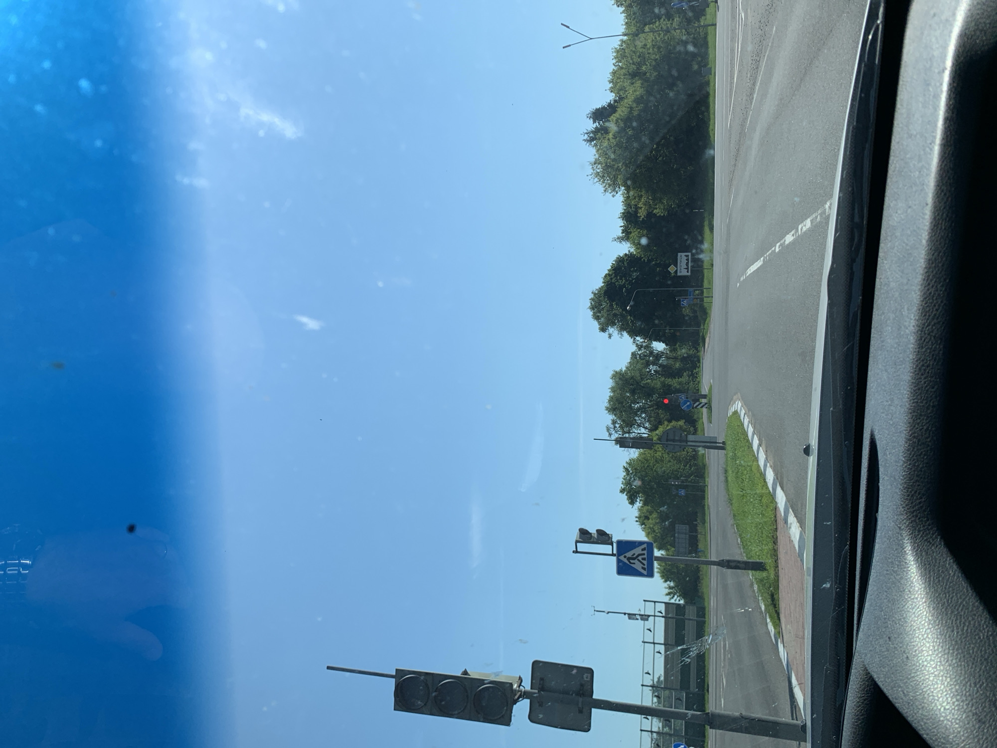 The image shows a view from inside a vehicle looking out through the windshield at a road intersection on a clear, sunny day. The road is paved and marked with white lines. There are traffic lights, pedestrian crossing signs, and trees visible. A sign in the distance appears to be a town or village name, "Шерешевцы". The foreground shows the dashboard of the vehicle.