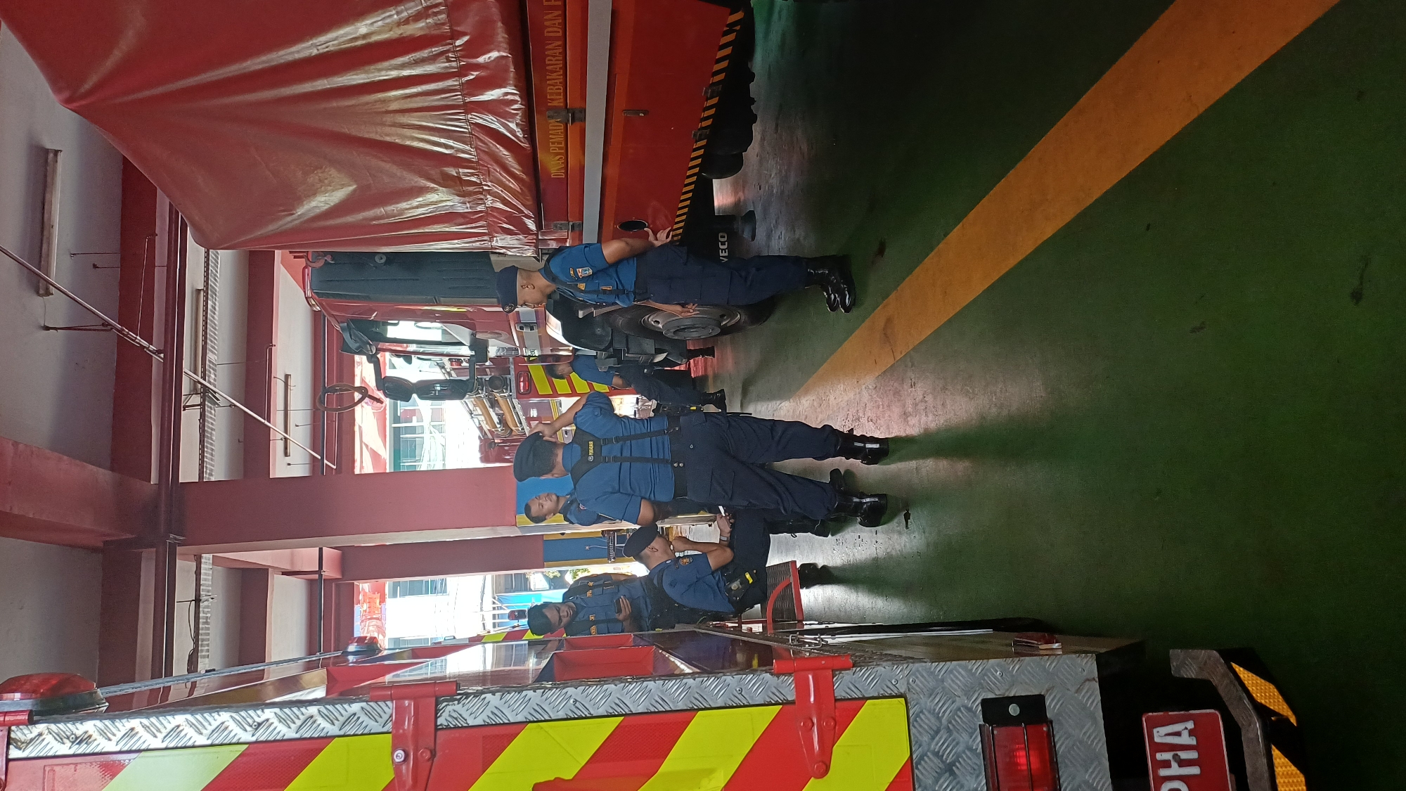 The image depicts a fire station interior with several firefighters in uniform gathered around two large red vehicles. One vehicle, on the left, is a fire truck with reflective yellow and red stripes. The other vehicle, on the right, is a larger truck with a red tarp covering its cargo area and text reading "DINAS PEMADAM KEBAKARAN DAN PEN". Several firefighters are standing and talking, while one is seated on a step, looking at a mobile phone. The floor is green with a yellow line marking a path. The lighting suggests an indoor environment, possibly a garage or bay area of the fire station. The context implies a routine gathering or briefing among the firefighters. The presence of fire trucks and uniforms indicates this is an operational fire station.