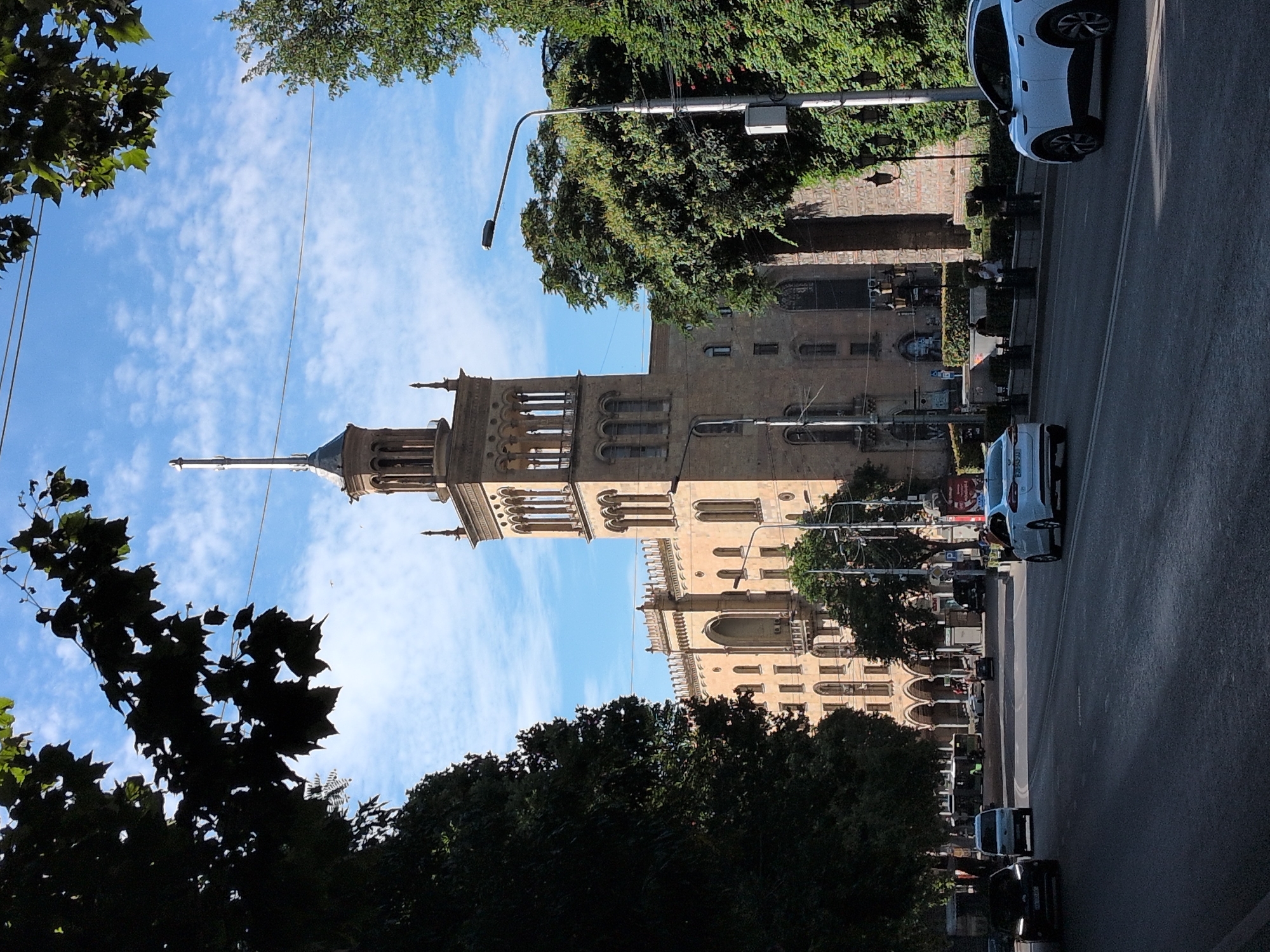 The image shows a street scene in Tbilisi, Georgia, with a prominent building featuring a tall, ornate tower. The building appears to be made of light-colored stone and has arched windows and decorative elements. Trees with lush green foliage frame the building and the street. Several cars are visible on the road, including a white SUV and a white sedan. The sky is blue with scattered white clouds. The scene conveys a sense of urban activity on a sunny day.