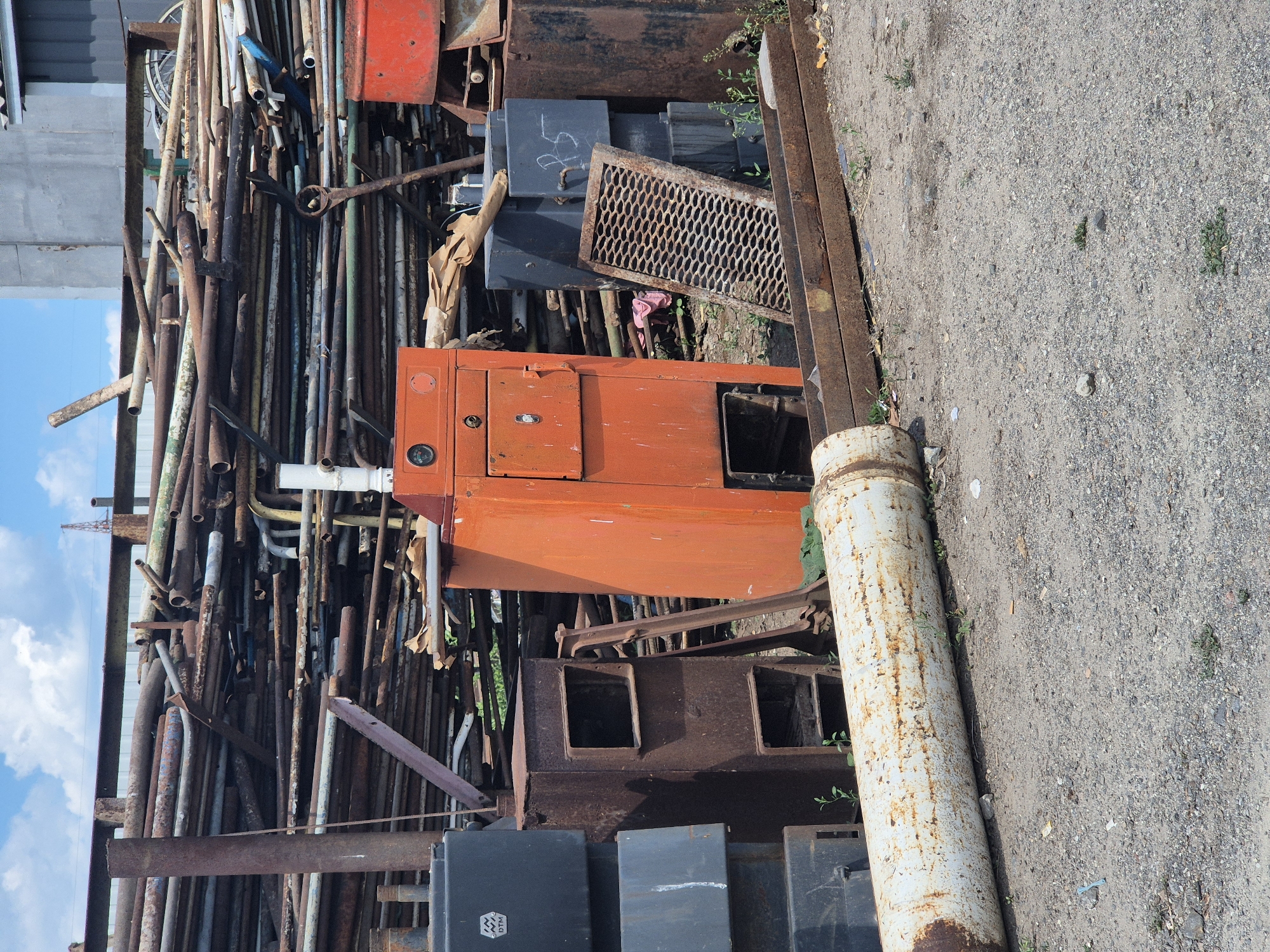 The image depicts an outdoor scene at what appears to be a scrapyard or industrial storage area in Pidhaitsi, Ukraine. In the foreground, a large, white, rusty pipe lies horizontally on the ground. Behind it, a collection of various metal objects are visible, including dark grey rectangular containers stacked on the left, a rusty, box-like structure with openings, and an orange industrial cabinet or boiler with a dial and a door. To the right of the orange cabinet, a weathered metal grate leans against a dark rectangular object. In the background, a massive pile of assorted metal pipes, some rusty and others painted in various colors including green and blue, are stacked high against a building structure. The ground is covered in gravel and dirt, with sparse green vegetation growing in places. The sky above is partly cloudy with a blue sky.