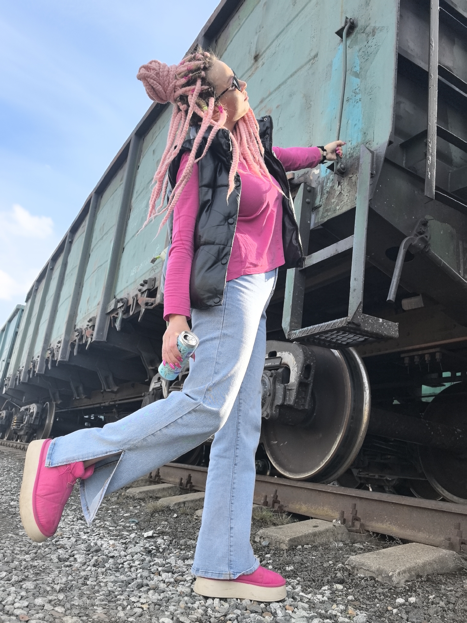 A woman with pink braided hair stands on a gravel surface near a train car in Chaikovsky, Russia. She wears a pink long-sleeved shirt, a black vest, blue jeans with slits at the ankles, and pink platform shoes. In her left hand, she holds a blue and silver can. She points towards the train car with her right hand. The train car is green and black, and sits on a railway track made of rusted metal and concrete blocks. The sky is blue with some clouds.
