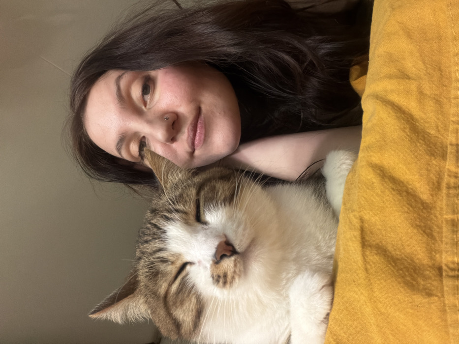 An indoor scene in the city of Chaikovsky, Russia, depicts a female individual and a domestic cat positioned in close proximity. The female, visible on the right, has long dark brown hair and fair skin. She wears a silver stud piercing in her right nostril and her right cheek rests on her right hand. Her eyes are open, directed towards the viewer, and her lips form a slight, closed smile. The cat, a tabby-and-white feline, occupies the left side of the frame. Its eyes are closed, and its head is slightly tilted, indicating a state of rest or contentment. A textured, crumpled yellow fabric, possibly a blanket or pillow, is present at the bottom of the frame, underneath the cat and extending towards the female. The background is an unadorned, light-colored wall or interior surface.