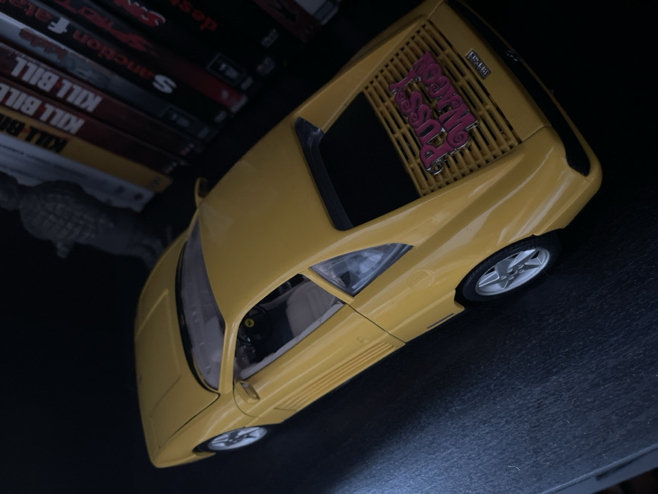 Within a dimly lit interior in Plaisir, France, a yellow die-cast model car, resembling a Ferrari 348, is positioned on a dark, flat surface. The model features a detailed interior with light-colored seating and a visible steering wheel. Its rear engine cover, characterized by horizontal slats, bears a prominent pink emblem inscribed with the words 'Pussy Wagon'. The vehicle is equipped with silver, star-spoke wheels. In the background, to the upper left, a blurry, dark grey statuette of a standing human figure is partially visible. To the right, a row of DVD cases or books with dark spines is arranged vertically, displaying titles including 'KILL BILL' (repeated) and 'Sanction'.