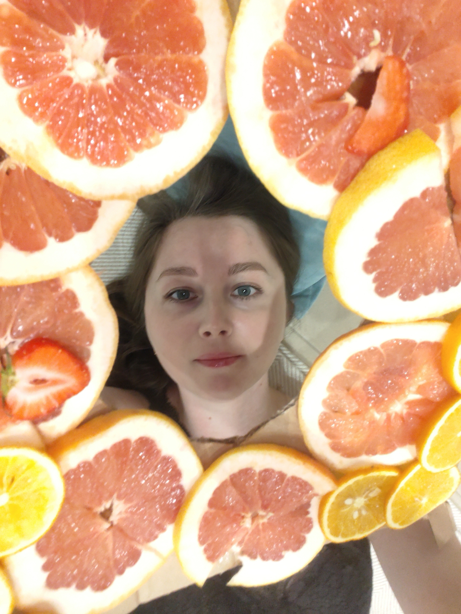 A selfie of a Caucasian woman with light brown hair and blue eyes, surrounded by slices of grapefruit and orange. One grapefruit slice contains a strawberry slice. The background appears to be a striped sheet or blanket. The location is unknown.
