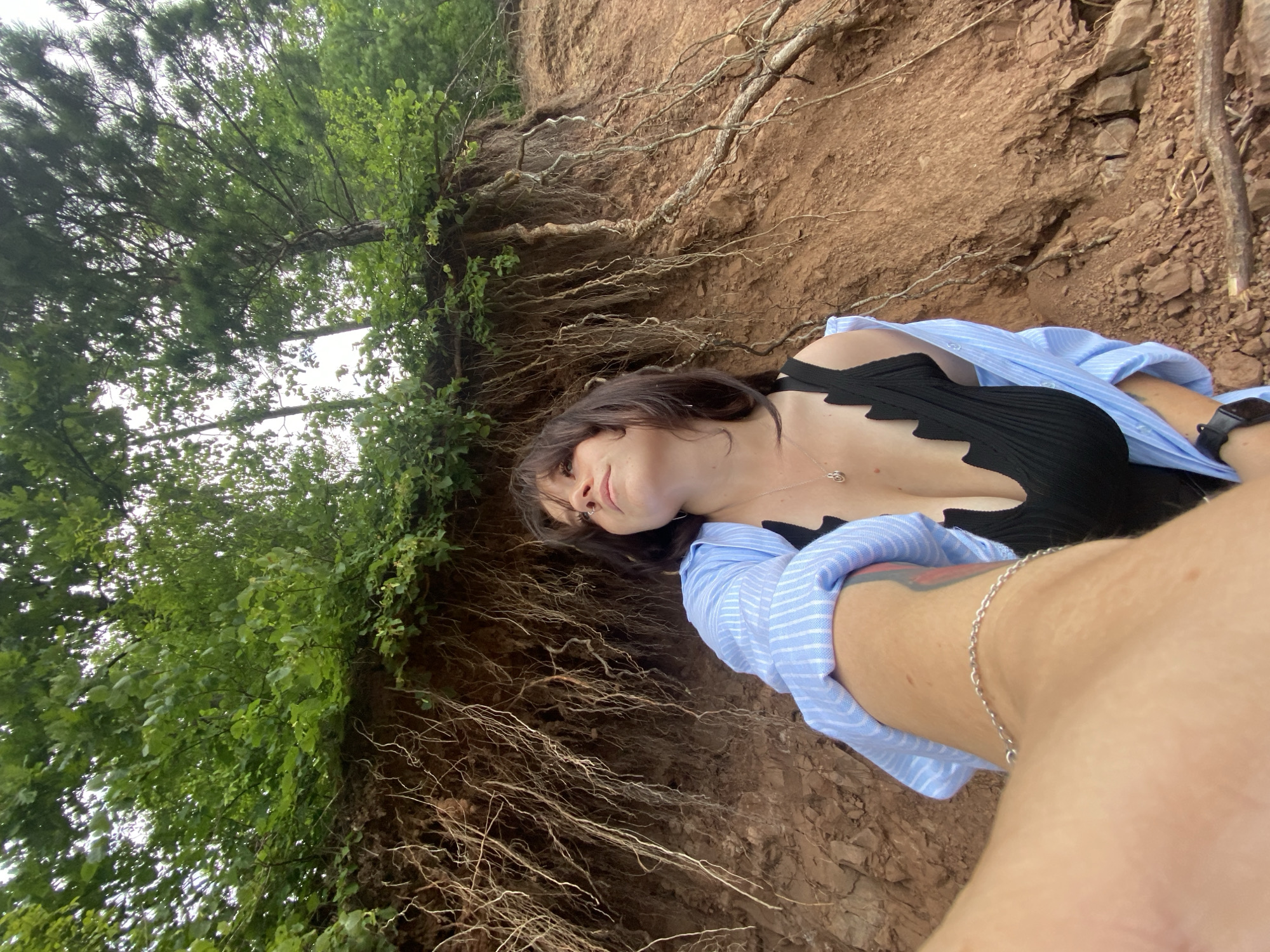 A low-angle, wide shot captures a young woman posing on a steep, eroded embankment with exposed tree roots. She is wearing a black, ribbed swimsuit with a zig-zag neckline and a light blue and white striped button-down shirt, worn open. A silver chain necklace with a small pendant is visible. Her hair is dark brown and styled with bangs. The embankment is composed of reddish-brown soil with numerous dangling roots and scattered small rocks and debris. Lush green foliage and trees line the top of the embankment, with a cloudy sky overhead. The lower portion of the image is dominated by a person's arm and shoulder, holding what appears to be a camera or phone, suggesting a selfie is being taken. The location is identified as Noviy, Russia.