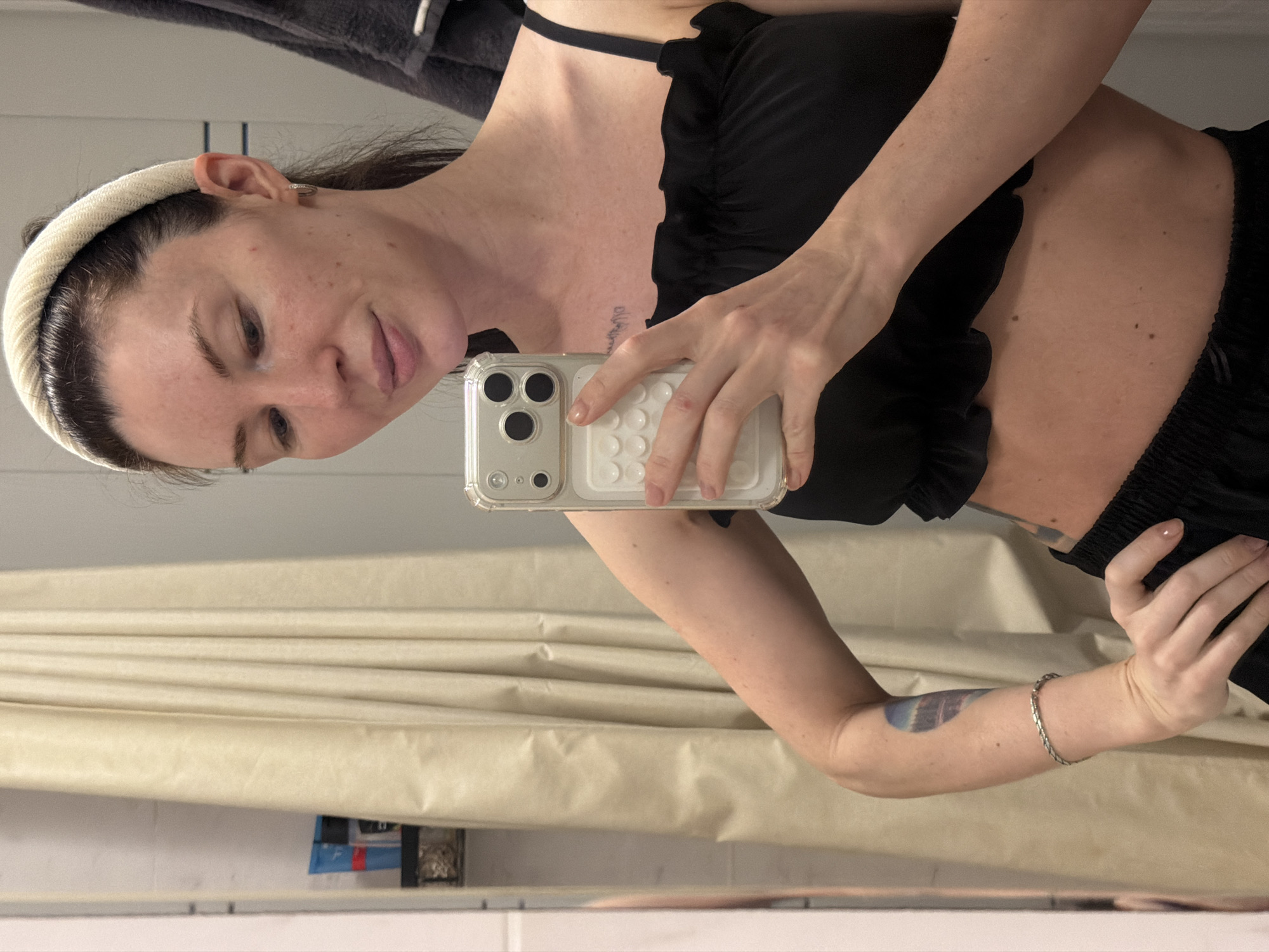 A woman takes a selfie in a bathroom with a beige curtain behind her. She is wearing a black top and a white headband. She has a tattoo on her left arm. The location is not discernible in the image, so it cannot be stated that the image was taken in Perm, Russia.