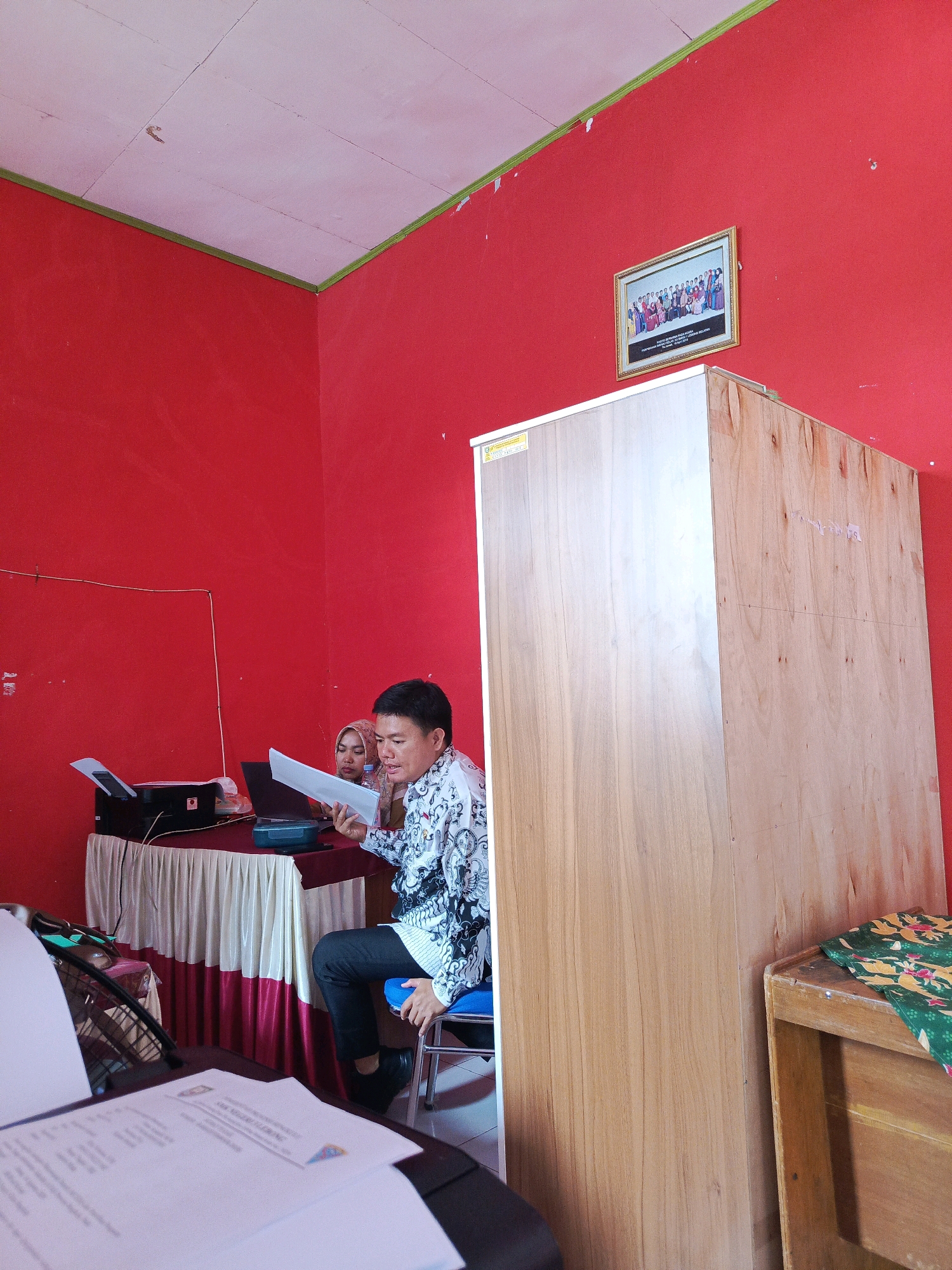 The image depicts an indoor scene with bright red walls and a white ceiling. In the foreground, a man in a patterned shirt and dark pants is seated on a blue chair, holding and looking at papers. To his left, a woman wearing a hijab is also looking at papers, positioned behind a table covered with a red and white striped cloth. On the table, a printer and a laptop are visible. To the right of the man, a tall, light-colored wooden cabinet stands against the wall. A framed picture of a group of people is hanging on the red wall above the cabinet. The scene appears to be a meeting or presentation taking place in an office or classroom. There is no direct indication of the city of Tik Jeniak, Indonesia, within the image itself.