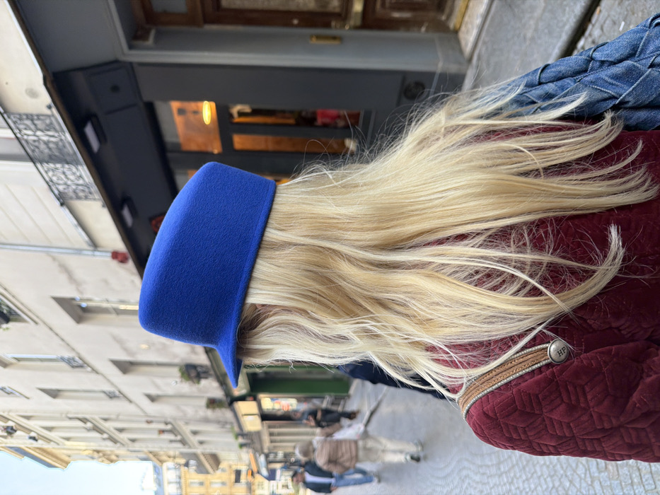 The image captures the back of an individual walking down a cobbled street in Paris, France, during daylight hours. The person has long, wavy blonde hair and wears a distinctive royal blue felt hat with a flat top and short brim. They are dressed in a maroon quilted jacket, on the left shoulder of which rests a tan leather strap featuring a circular silver button embossed with "HP." A blue woven bag is visible slung over the right shoulder. The background features characteristic light-colored Haussmannian buildings with multiple floors, dark window frames, and ornate wrought-iron balconies. Ground-level storefronts, including one with a dark grey facade and a prominent wooden door, line the street. Several blurred figures of other pedestrians are observable in the mid-ground, indicating a populated urban environment.