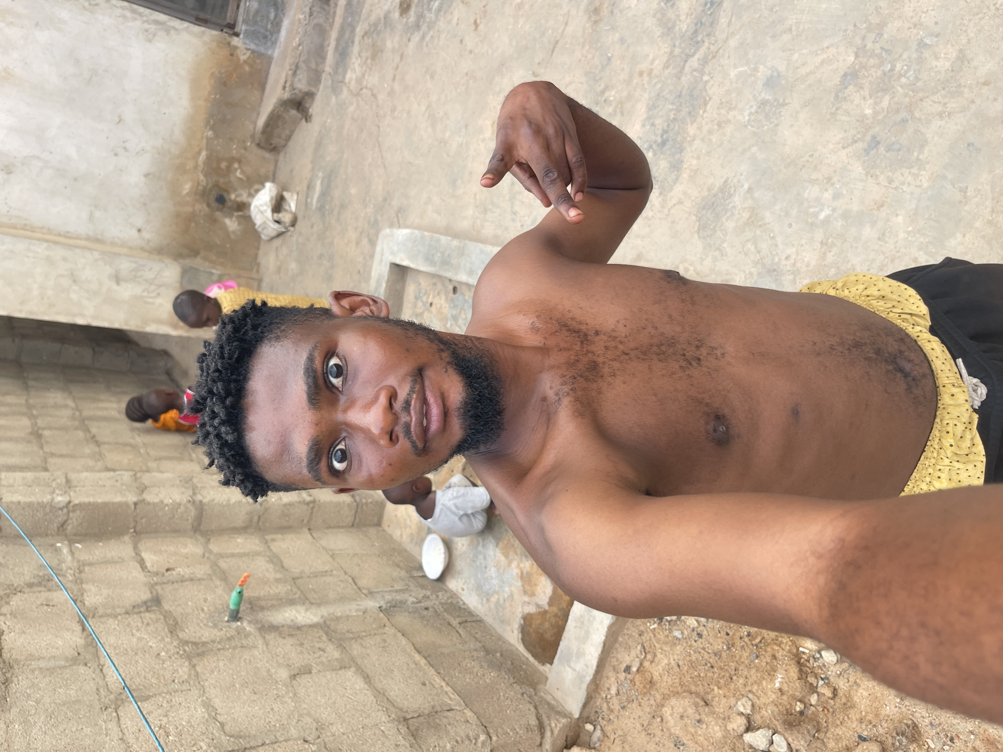 A shirtless man with dark hair and a beard takes a selfie in Unguwar Gabas, Nigeria. He wears shorts with a yellow and black pattern. Two children are visible in the background, one lying down and one sitting. The setting appears to be an outdoor concrete area. A blue wire stretches across the lower-left corner of the frame. A green and orange object is stuck in the ground.