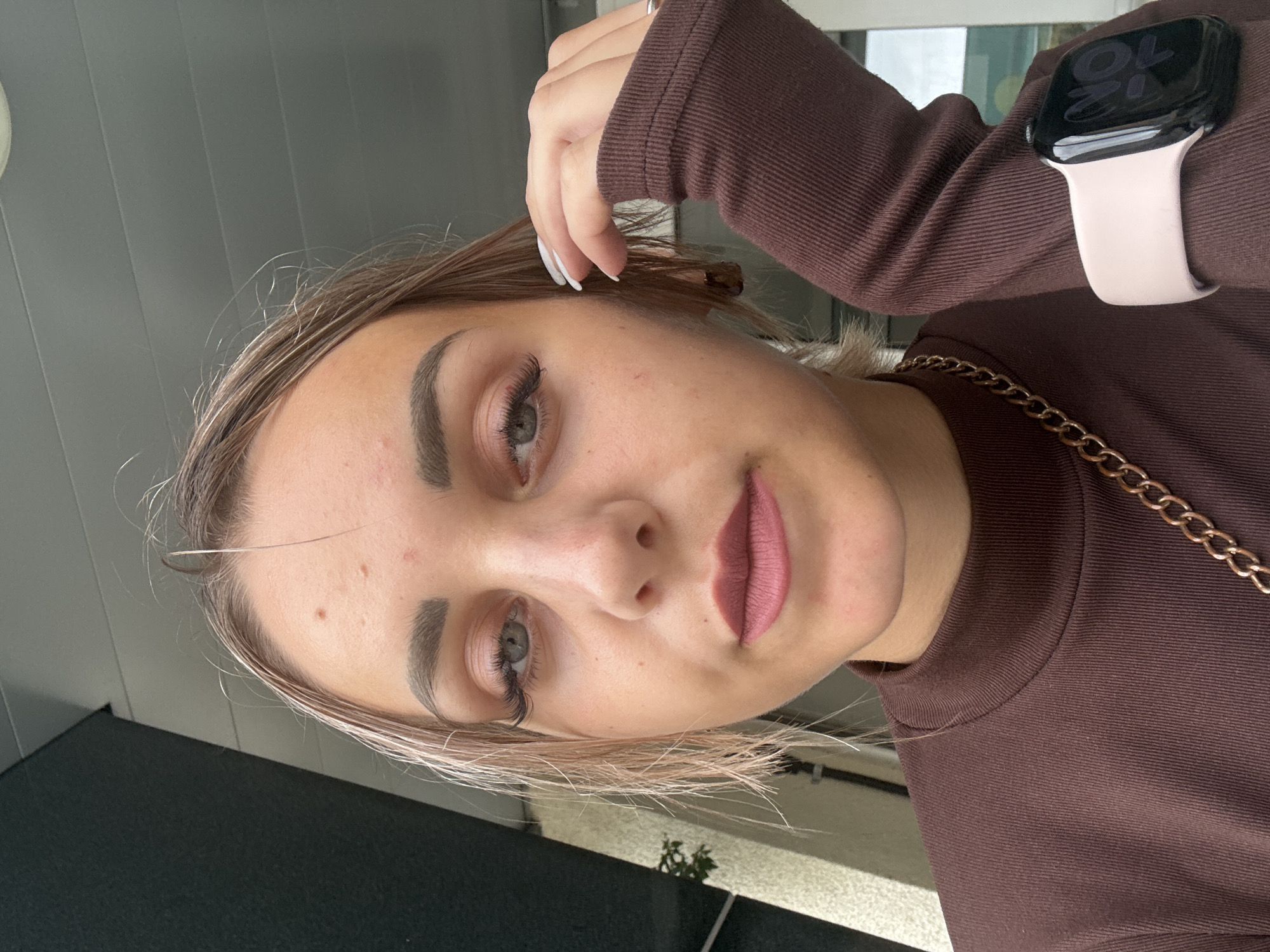 A young woman with light brown hair and light skin is pictured in a close-up selfie. She is wearing a dark brown, high-necked, ribbed shirt with a gold chain necklace visible at the collar. Her left hand is touching her hair near her temple. She has on dramatic false eyelashes and matte pink lipstick. A light pink Apple Watch is worn on her right wrist. The background suggests an outdoor setting with some architectural elements, possibly a building facade or overhang. There are no discernible landmarks or environmental cues that would specifically place this image in Yuzhno-Sakhalinsk, Russia.