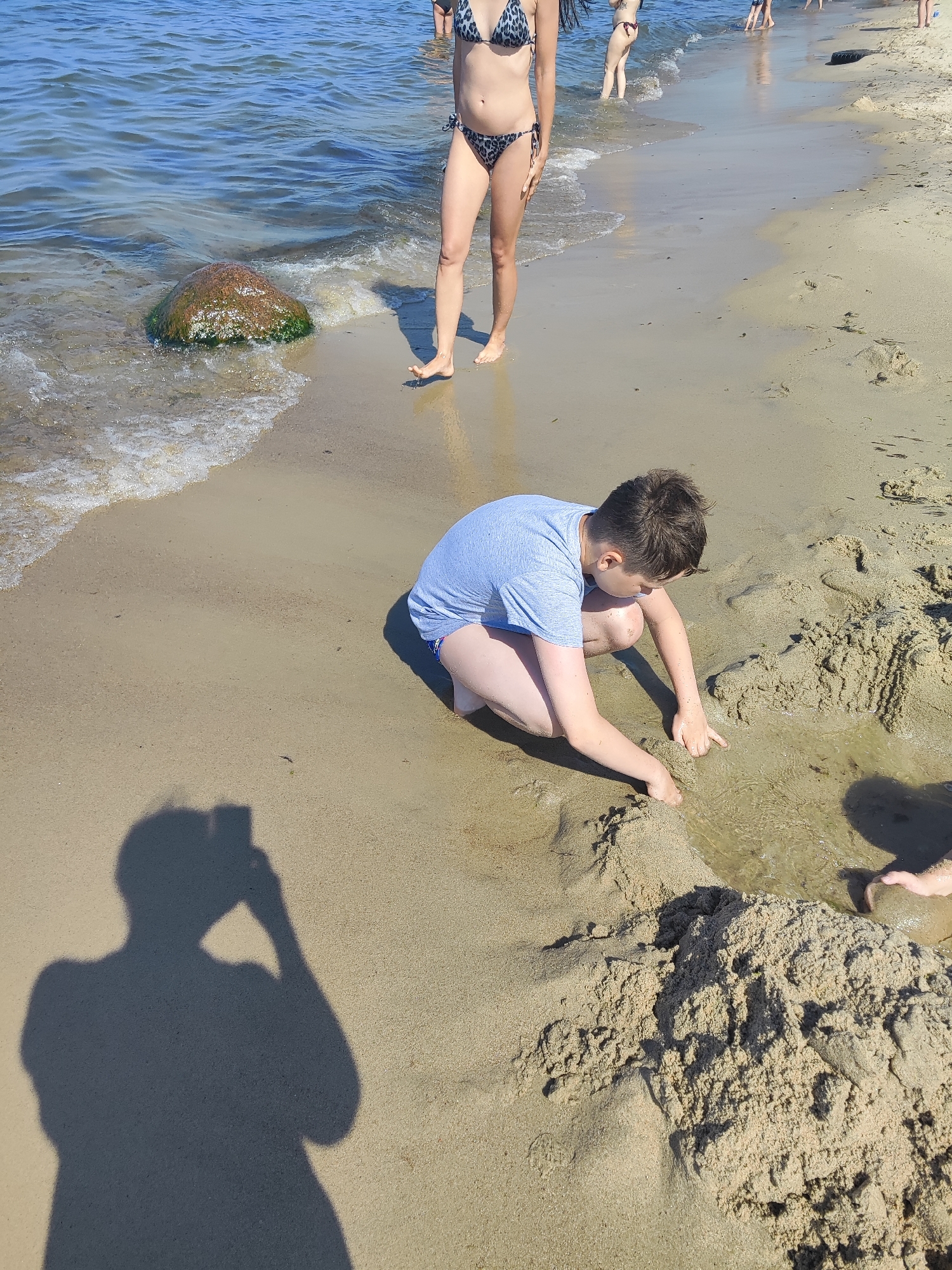 The image captures a scene on the sandy beach of Pionersky, Russia, with the Baltic Sea in the background. In the foreground, a young person wearing a light blue t-shirt and swim shorts is crouching on the sand, actively playing in a shallow pool of water. Their hands are in the water, and they appear to be digging or manipulating the sand. To the left, the shadow of a person holding a camera is cast on the sand, indicating the photographer's presence. Further back, two individuals in bikinis are standing by the water's edge. The beach is composed of light-colored sand, and the water is calm with gentle waves lapping at the shore. A large, moss-covered rock is partially submerged in the water near the shoreline. The overall atmosphere is that of a sunny day at the beach.