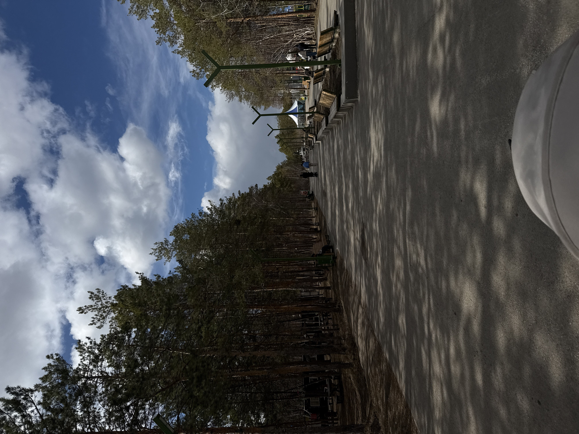 The image captures a pedestrian walkway in a park in Yakutsk, Russia. The walkway is paved and shaded by the shadows of trees, extending into the distance. The left side of the frame shows a dense line of tall pine trees. Along the right side of the walkway, there are benches and stylized green lampposts. In the distance, there are a few people walking along the path. The sky is visible above the trees, with a mix of blue sky and puffy white clouds. A partial view of a white object is visible in the lower right corner.