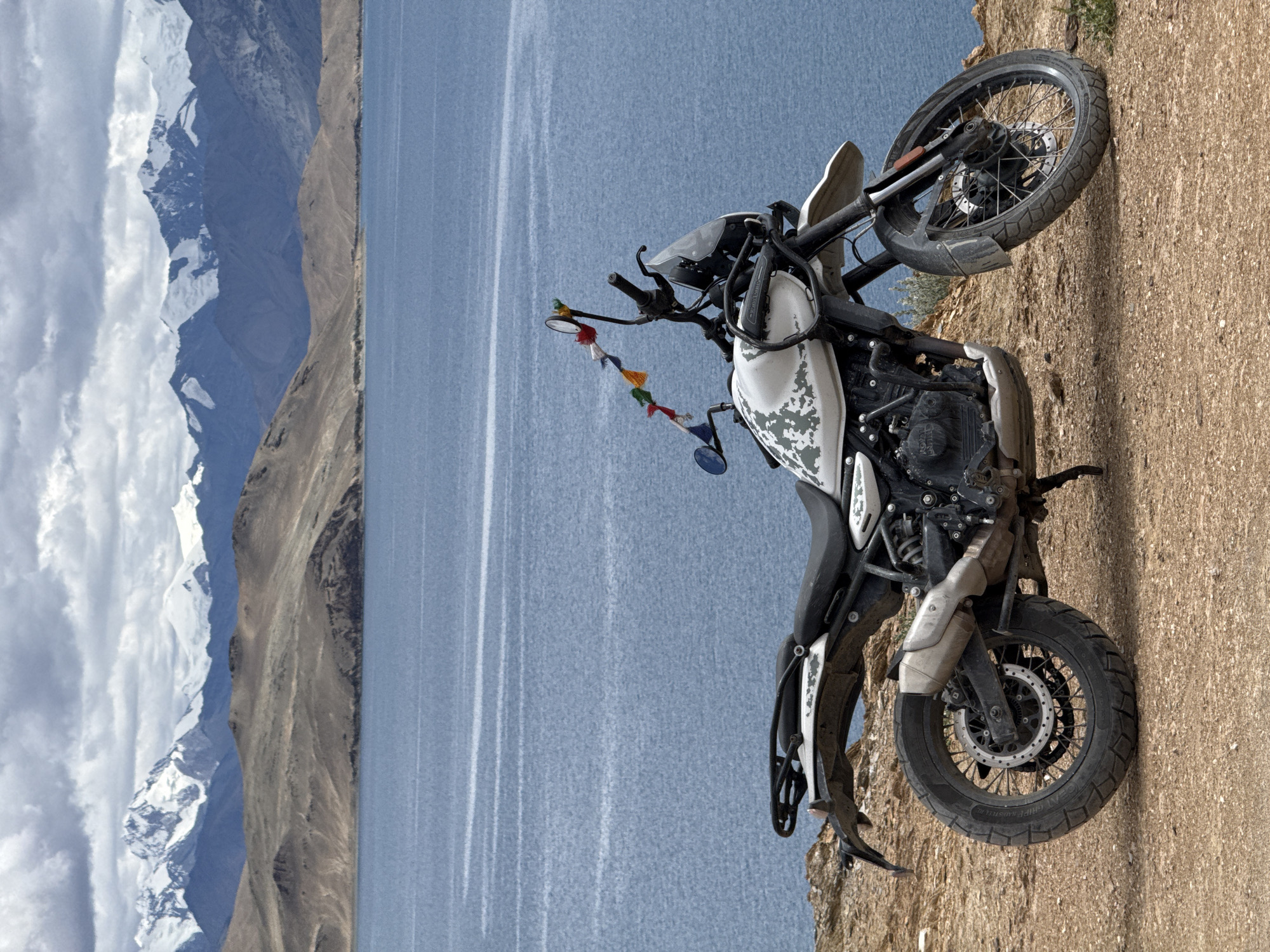 An off-road motorcycle is parked on a rocky outcrop overlooking a vast, calm blue body of water. The motorcycle is white with a camouflage pattern and prayer flags are attached to the handlebars. In the background, there are rolling, barren hills leading up to snow-capped mountains under a cloudy sky. The location is likely in the Himalayas, given the landscape and prayer flags, suggesting a remote, high-altitude area of India.