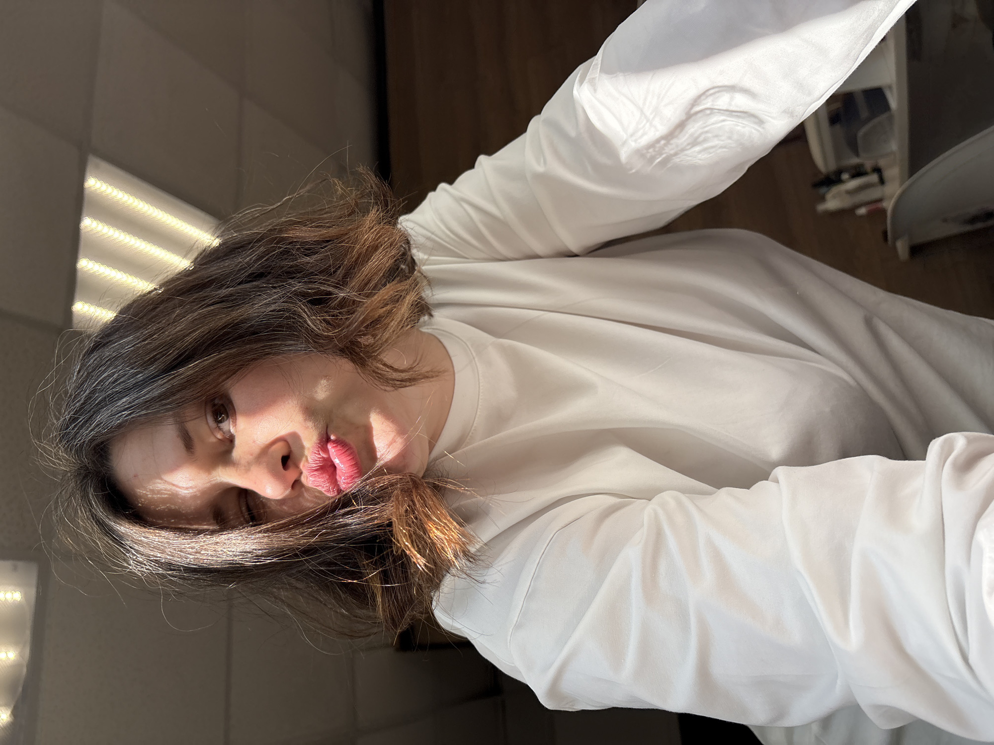 A woman with dark hair and red lips is facing down and to the left of the image, with light reflecting on her face and hair. She is wearing a long-sleeved white shirt. The background includes a dark surface, possibly a wall or floor, and a light source from the upper left of the image. The exact location in Chaikovsky, Russia is not identifiable.