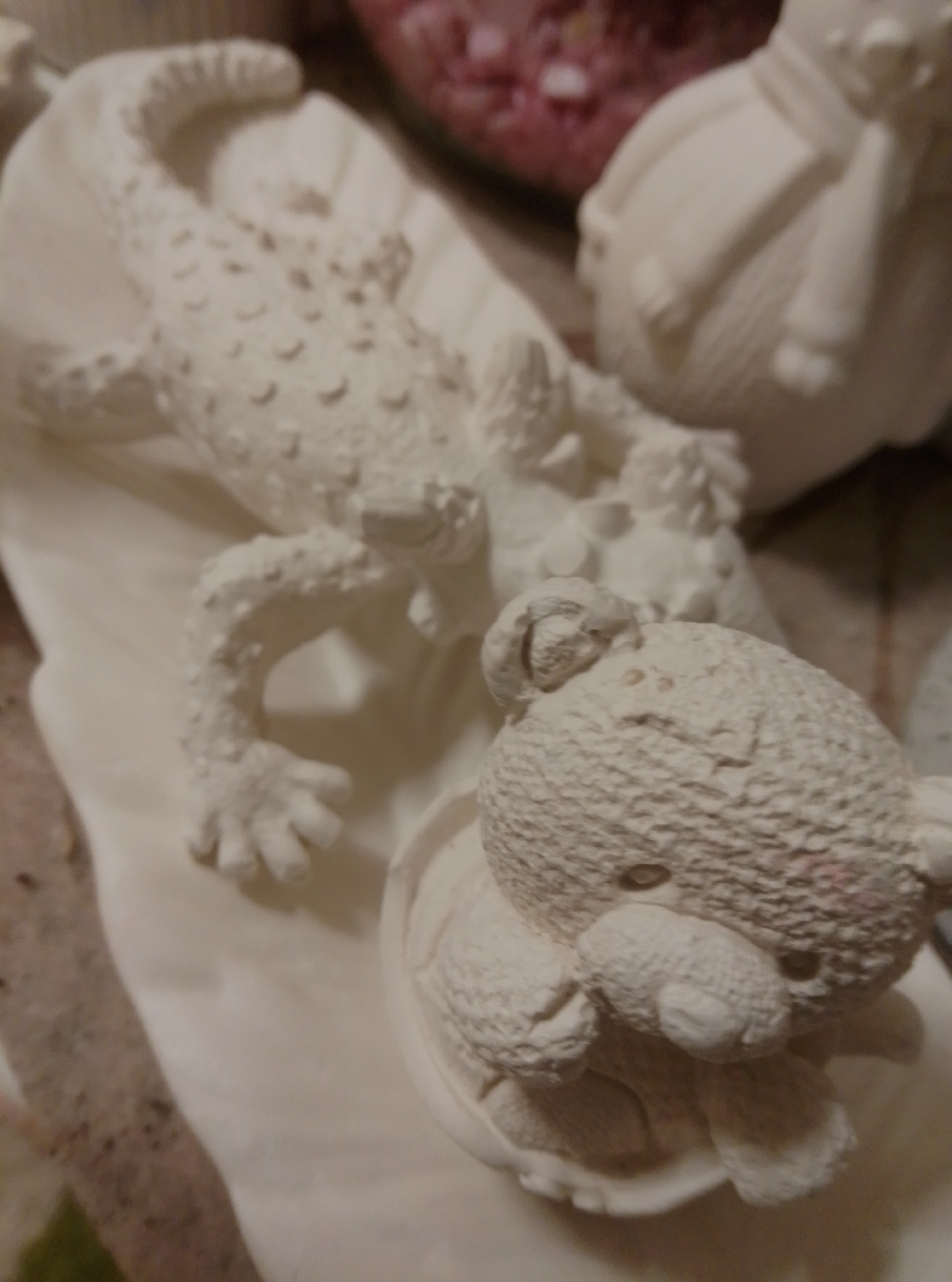 The image shows a close-up, slightly blurred view of several plaster figures. In the foreground, a teddy bear figure is nestled in a white bowl-like object. The teddy bear has textured fur, visible eyes, a snout, and paws. Behind the teddy bear, a longer, scaly figure resembling a lizard or alligator lies on a white surface. This figure has distinct legs, feet, and a textured body. In the background, another plaster figure, possibly a bird or a round character, is partially visible. The overall scene is composed of white, unpainted plaster figures, suggesting they are either finished products or in the process of being decorated. The background appears to be a surface with some pinkish, blurry objects, possibly other decorations or materials. The image does not provide any information about the location within the city of Wedel, Germany, or any specific context related to its placement there.