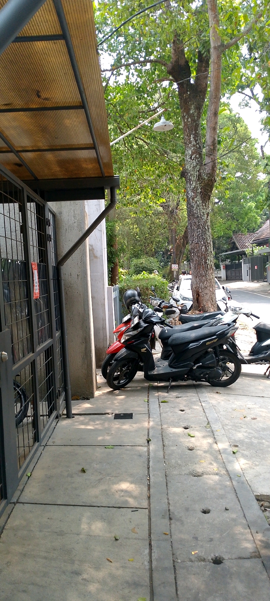The image shows a street scene in Bandung City, Indonesia, with a focus on the sidewalk and parked motorcycles. A covered walkway with a translucent corrugated roof supported by metal beams extends from a building on the left, creating a shaded area over the sidewalk. A metal gate with a grid pattern is part of the building's facade. Two motorbikes are parked parallel to the building on the concrete sidewalk. The front motorbike is red and black, with a black helmet on its seat. The motorbike behind it is black. Further down the street, another white motorbike is visible, along with a white car. A large tree with rough bark stands next to the street, its branches reaching over the scene. The environment is urban, with buildings and residential roofs visible in the background. The ground is paved with concrete slabs, and fallen leaves are scattered on the surface. The scene appears to be captured during the daytime, with sunlight filtering through the trees, casting shadows on the ground.