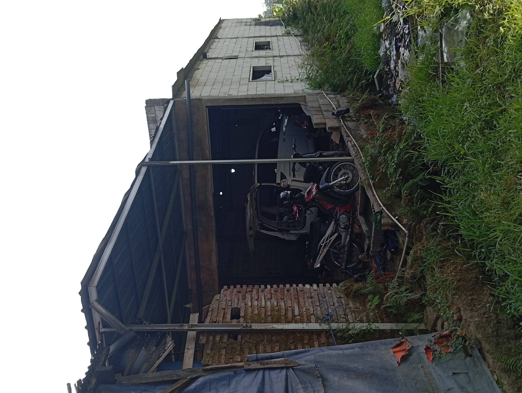 The image depicts an outdoor scene in Lemahbang, Indonesia, featuring a partially constructed or dilapidated building with a metal roof and walls made of brick and concrete blocks. The structure appears to be an open-air workshop or garage. Inside the structure, a white van with its rear doors open is parked, along with a motorcycle. Various tools and materials are visible within the workshop area, suggesting ongoing work or storage. In the foreground, lush green vegetation and some scattered debris are present. The sky is visible at the top of the image, appearing bright and possibly overcast.