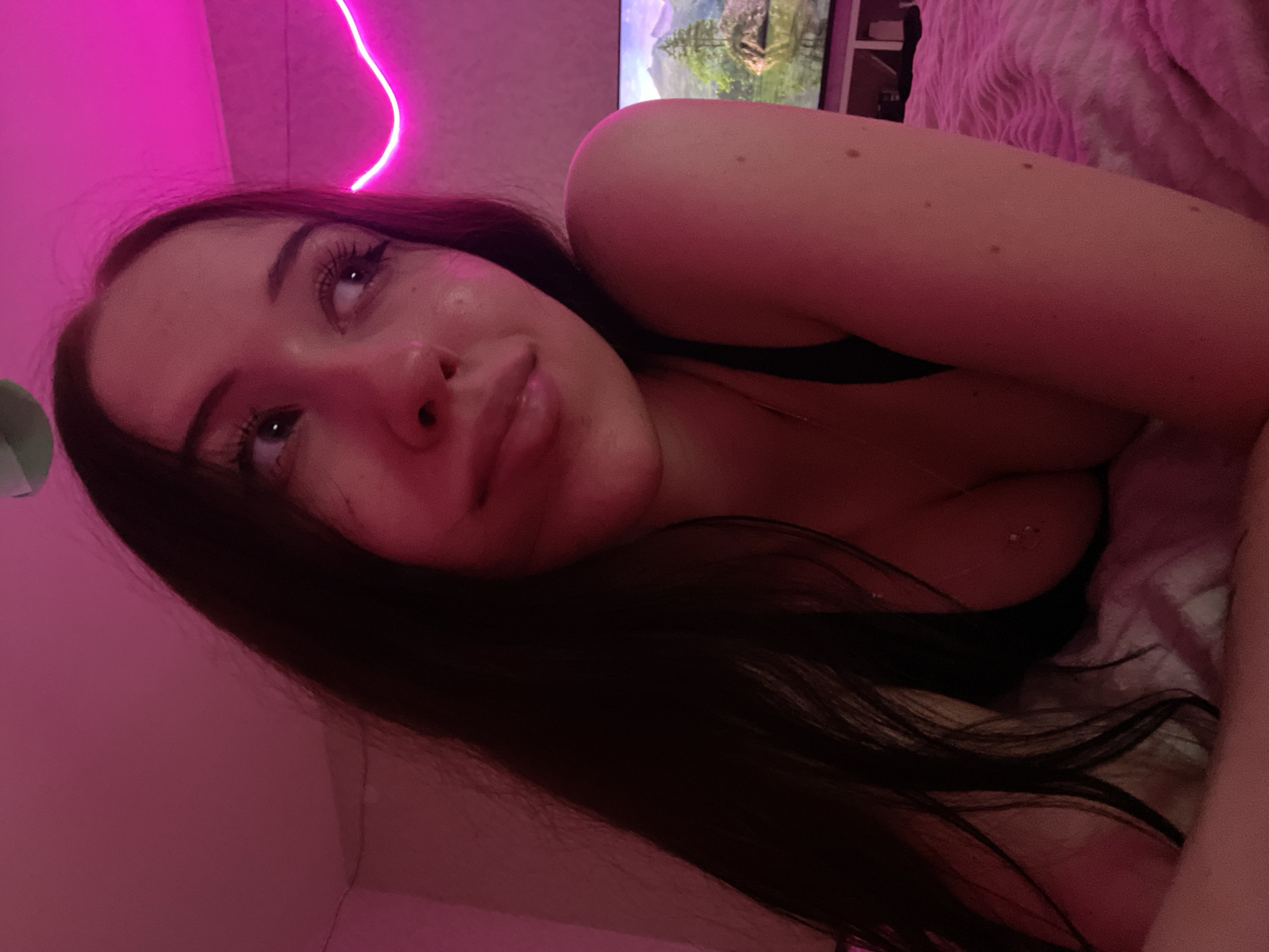 A young woman with dark hair and fair skin is lying down indoors, illuminated by pink lighting. She is wearing a black top that reveals her cleavage. In the background, a television screen displays a landscape image. The overall environment suggests a bedroom setting. The image does not provide any information that could be used to determine the location within Votkinsk, Russia.