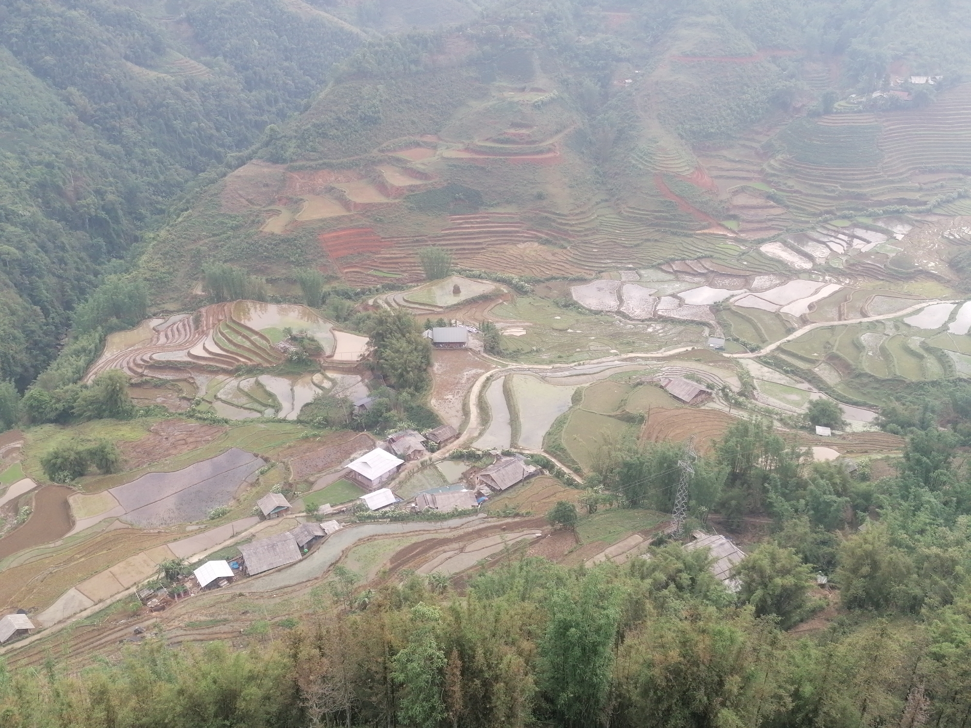 The image shows an aerial view of the countryside in Sa Pa Ward, Vietnam. The landscape is dominated by terraced rice paddies on the hillsides. Some paddies are filled with water, while others are dry or show early growth of crops. A small village with houses with corrugated metal or thatched roofs is nestled among the terraces. Patches of trees and bamboo clumps are scattered throughout the area, and a few trails or dirt roads wind through the village and the paddies. The overall scene is hazy, with a soft, diffused light. An electrical transmission tower is also visible.