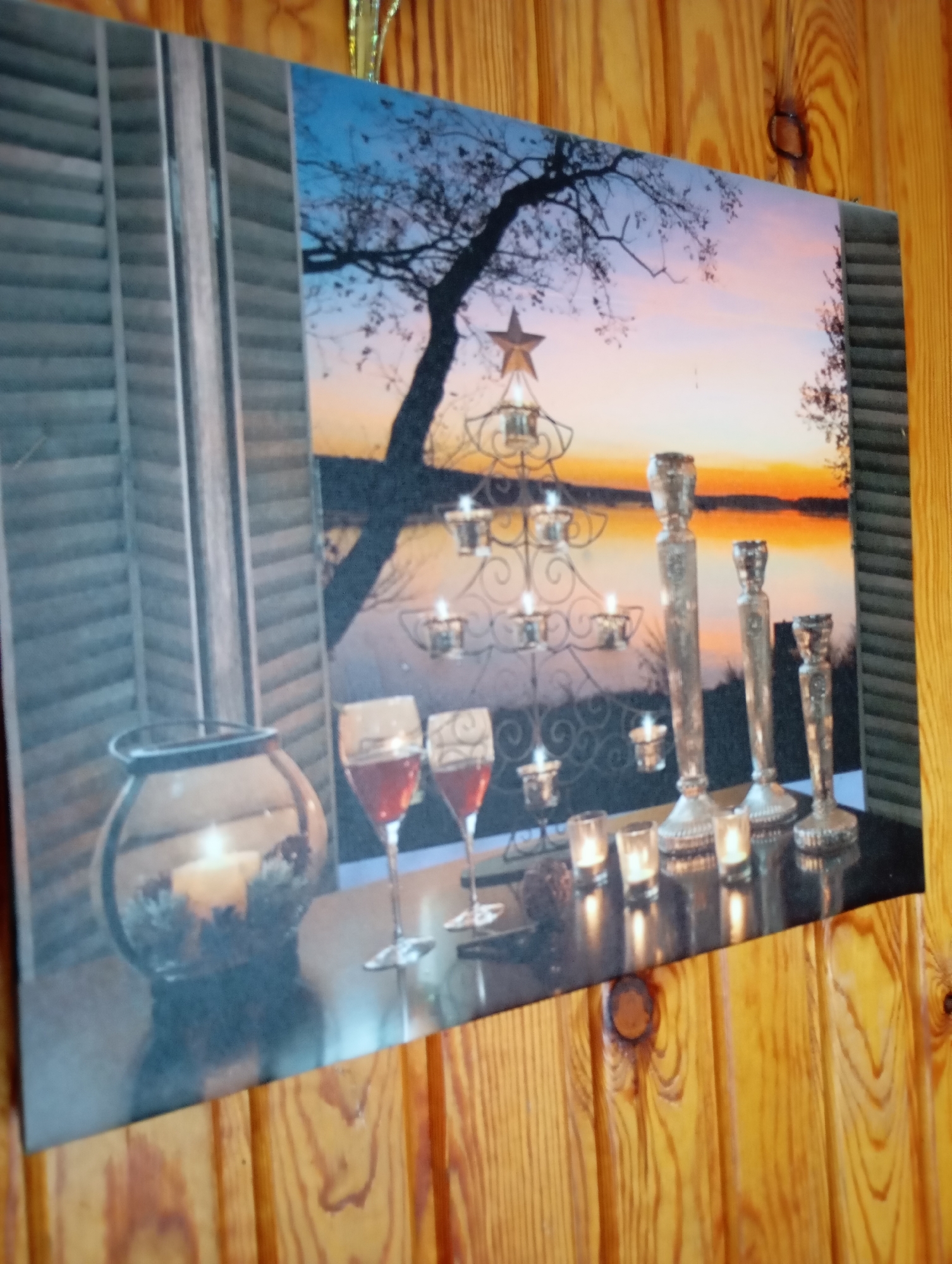 This is a photographic print on canvas depicting a still life scene set against a sunset landscape. The foreground features a dark, reflective surface adorned with several objects: a glass hurricane lantern containing a lit candle, two wine glasses filled with red wine, and a decorative candelabra holding multiple lit tea lights. To the right of the candelabra are three ornate silver-toned candle holders of varying heights, each with a lit candle.
The background of the print shows a twilight sky with hues of orange, pink, and blue, reflecting on a body of water. A silhouetted tree with bare branches and a star ornament at its top is visible. To the left of the scene, there is a depiction of interior shutters.
The canvas is mounted on a wooden wall, the texture of which is visible around the edges of the print. The lighting suggests an indoor setting where the print is displayed. The image does not contain any people or animals, and no specific location in the city of Льниха, Russia can be identified from the visual information presented.