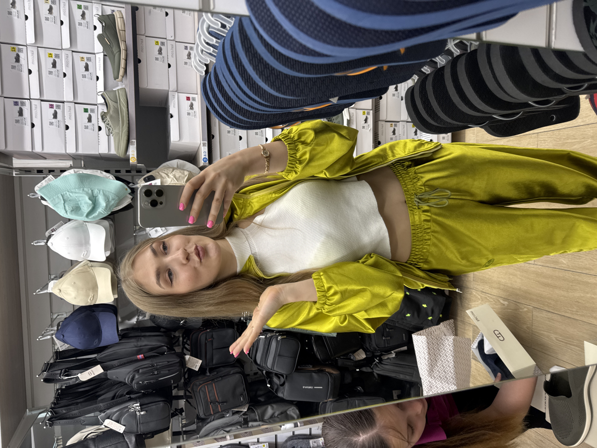 A young Caucasian female is taking a selfie in a retail store, posing with her lips pursed. She is wearing a lime green satin tracksuit consisting of a zippered jacket and drawstring pants, and a white ribbed crop top. The store appears to sell bags, shoes, and hats, with shelves stocked with boxed footwear and displays of various bags and caps. In the background, other shoppers are visible in the reflection of a large mirror. The store environment suggests a casual shopping experience.