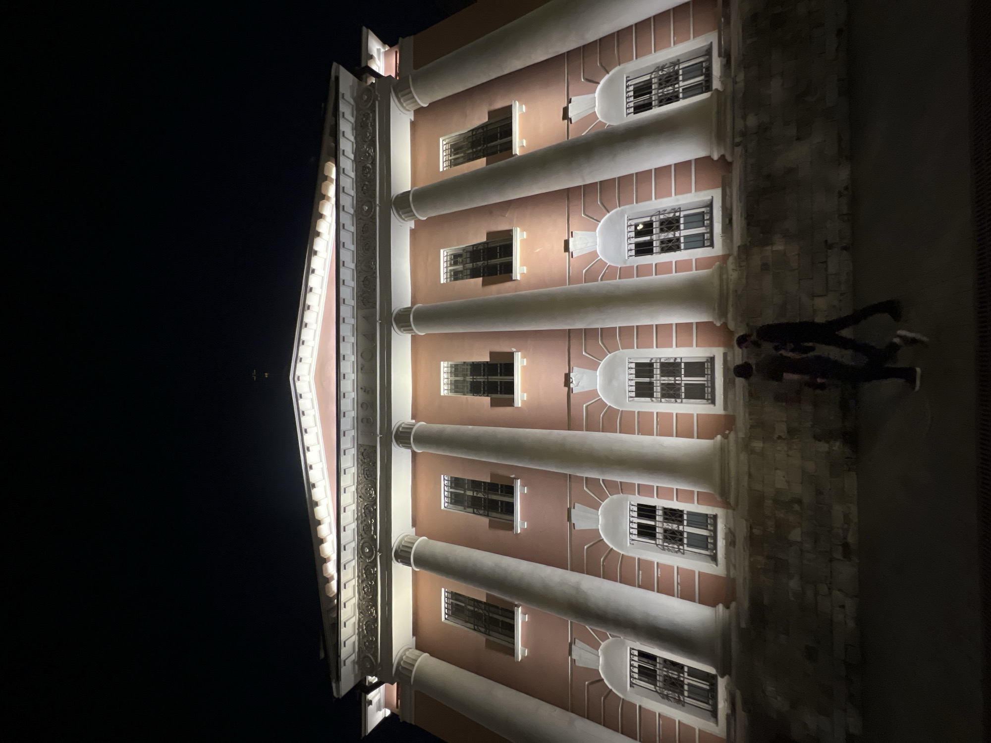 At night, a large pink building with white columns and windows is illuminated. Two people are walking in front of the building. The building appears to be an old-fashioned institution, possibly a museum or a government building, judging by its classical architecture. The windows have metal bars on them, suggesting a historical or security purpose. The street in front of the building is dark, with some lighting from the building itself casting shadows. The overall atmosphere is quiet and serene, typical of a city at night.