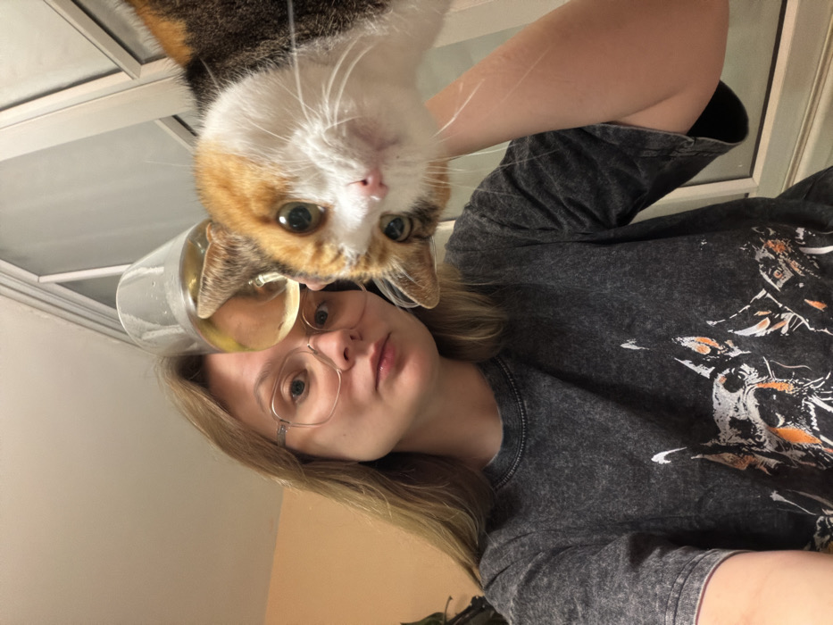 The image presents an indoor, high-angle, selfie-style view featuring a single human subject and a cat. A young adult female, with long blonde hair and clear-framed eyeglasses, occupies the left side of the frame, looking directly at the camera with a neutral expression. She wears a dark grey, possibly stone-washed, short-sleeved t-shirt featuring a graphic print of multiple dog heads, with some orange accents visible. To her right, a calico cat, predominantly white on its face with patches of orange and dark brown/black fur on its head and body, peers towards the camera with wide, green or amber eyes. A transparent drinking glass containing a light yellow liquid is positioned above the cat's head and near the woman's head, partially obscuring the cat's right ear. The background includes a plain, light orange-colored wall to the far left and a white-framed window or glass door on the right, through which an indistinct, pale outdoor environment or sky is visible. The internal environment appears well-lit. There are no identifiable landmarks or unique architectural features present in the image to confirm the specific location within Tel-Aviv, Israel.