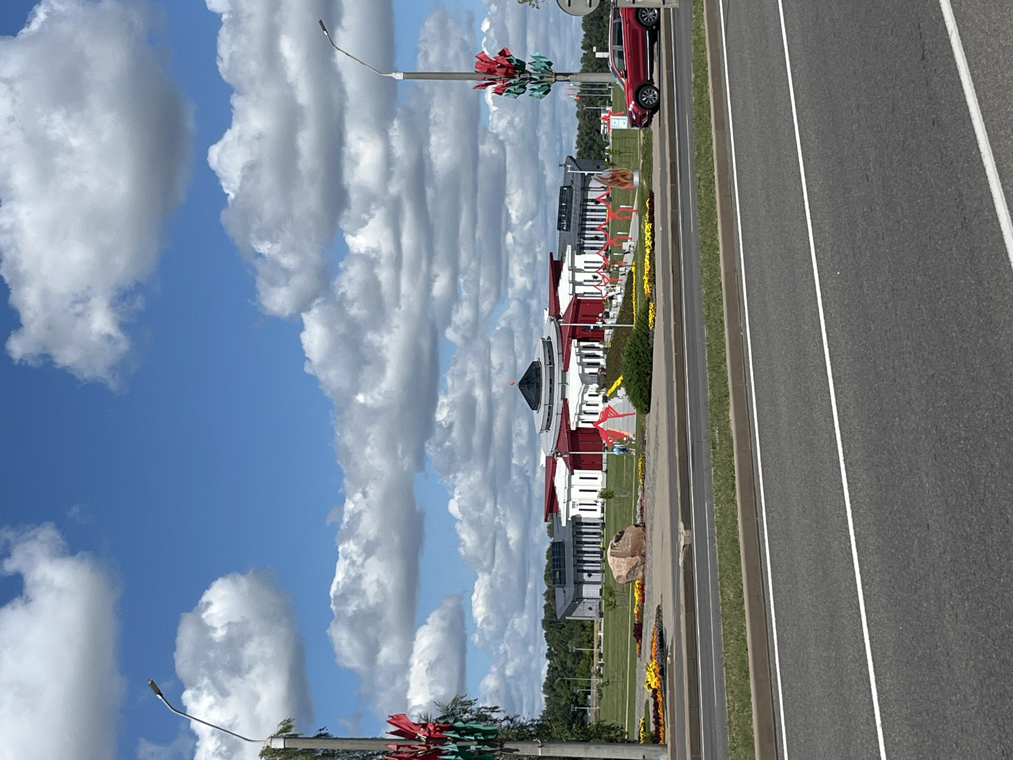 The image captures a view of the Khatyn Memorial Complex in Буйніцкі сельскі Савет, Belarus, on a cloudy day. In the foreground, a multi-lane asphalt road with white lane markings is visible. A strip of green grass and a concrete curb separate the road from the memorial grounds.
The memorial itself features several white buildings with red roofs and accents. Some of these buildings have rows of tall, narrow windows. A large, dark, domed structure with a pointed roof is also part of the complex, topped with a red spire. Decorative elements, including red ribbons and what appear to be flower displays, adorn the grounds. A large, rough-surfaced boulder is situated in the middle of a landscaped area with colorful flower beds. Two lampposts, decorated with what look like red and green artificial flowers or foliage, are positioned on the left and right sides of the scene. A bright red SUV is parked to the right of the memorial. In the distance, a line of green trees forms the horizon. The sky is a vibrant blue, dotted with large, fluffy white clouds. Two small figures, possibly visitors, are faintly visible walking on the memorial grounds.