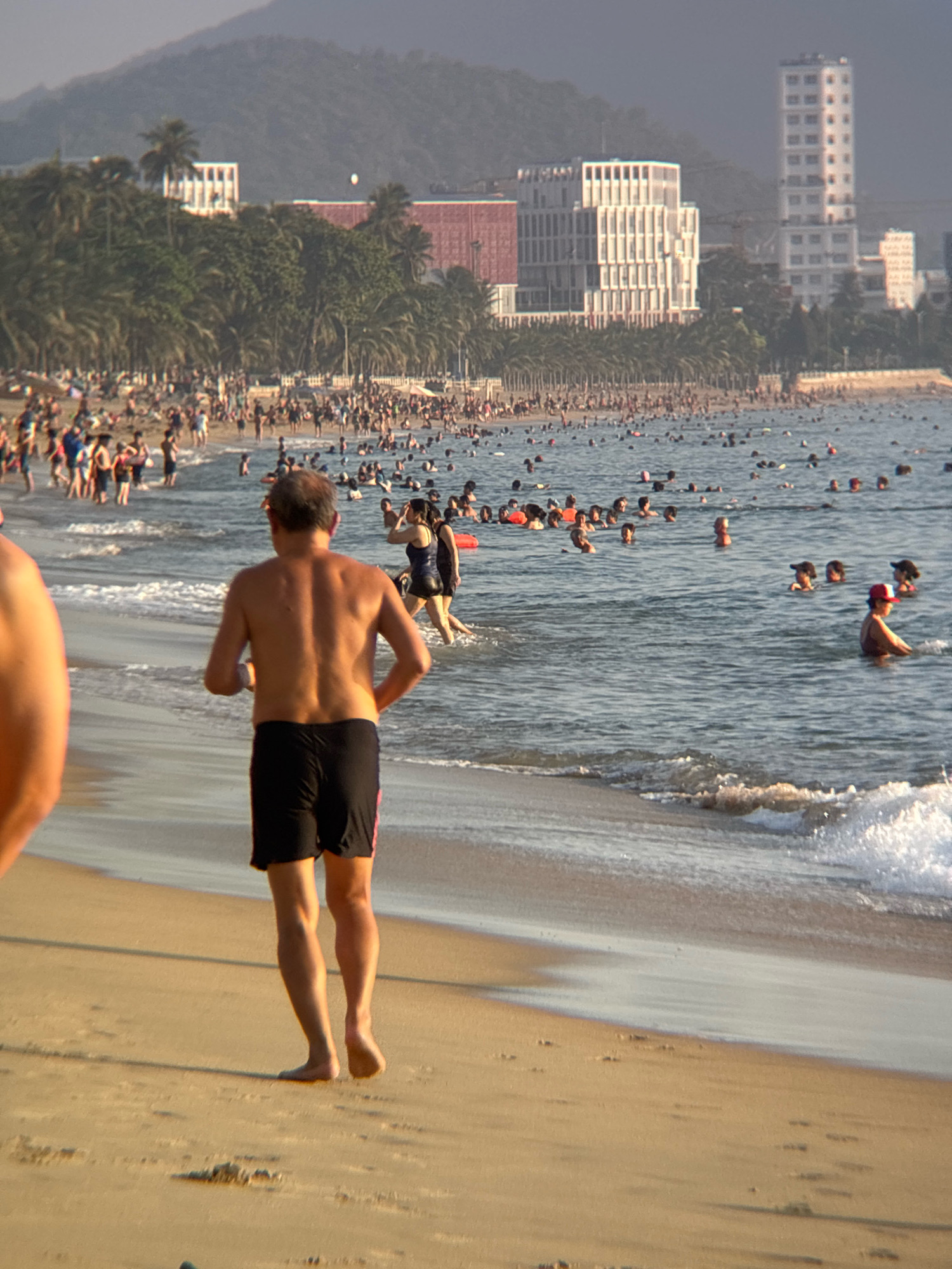 The image depicts a crowded beach in Nha Trang, Vietnam. The foreground shows a man in black swim shorts running on the sand. In the background, a vast expanse of the ocean is filled with numerous people swimming and wading. The shoreline is lined with palm trees and several modern buildings, including a tall white skyscraper and a multi-story structure with a distinct architectural style. The water is calm with gentle waves lapping the shore. The overall scene suggests a vibrant and busy recreational area on a sunny day.