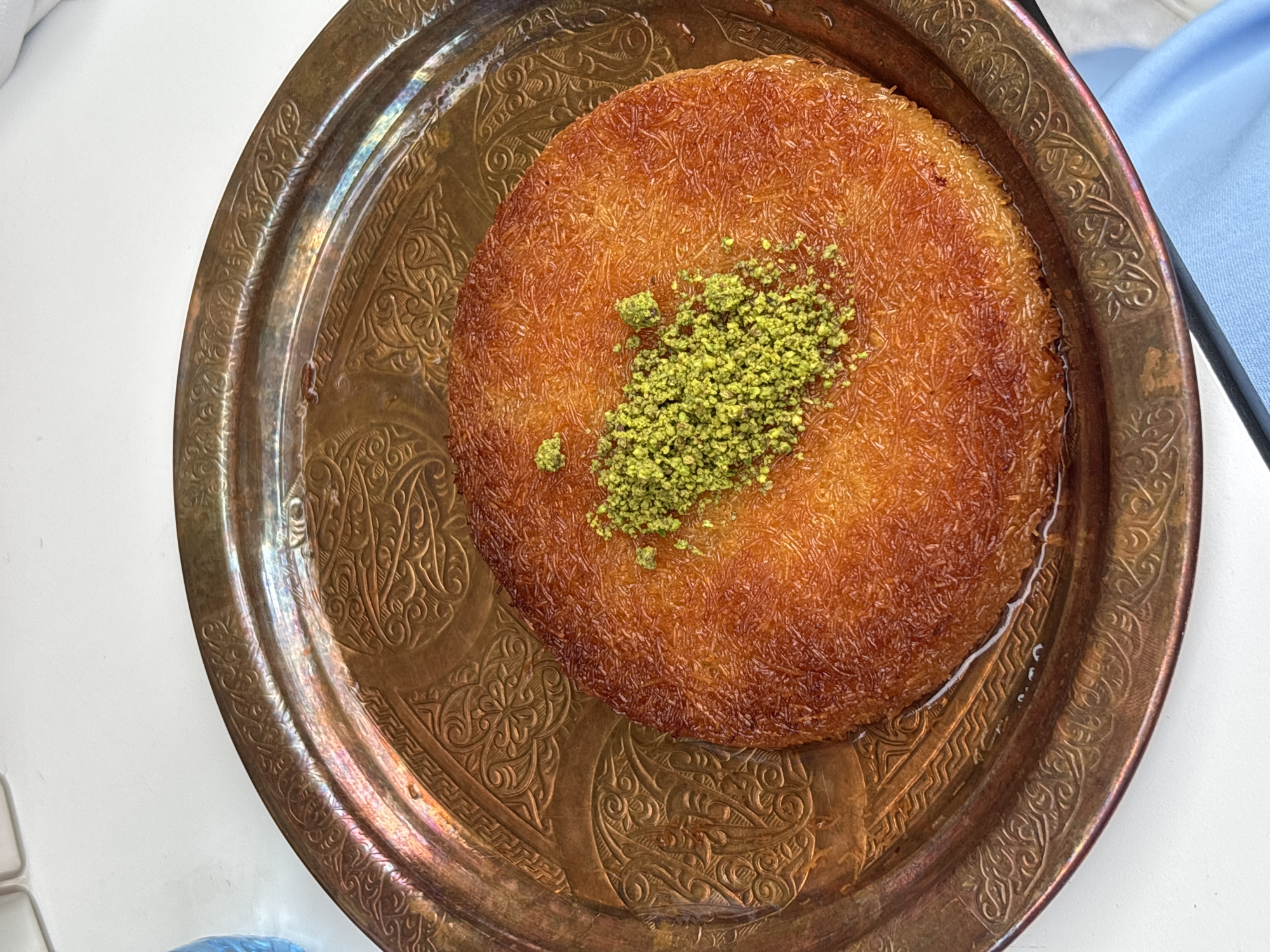 A top-down, medium-shot captures a dessert, possibly Kanafeh, served on a decorative bronze plate. The dessert is round, with a golden-brown, shredded wheat-like texture, topped with a small mound of green, possibly ground pistachios. A thin liquid, likely syrup, surrounds the dessert within the confines of the plate. The plate itself is round with an ornate, embossed design. The background is out of focus, but a white surface is visible underneath and a hint of light blue fabric is seen on the side. This dessert may be located in the city of Unknown, Unknown.