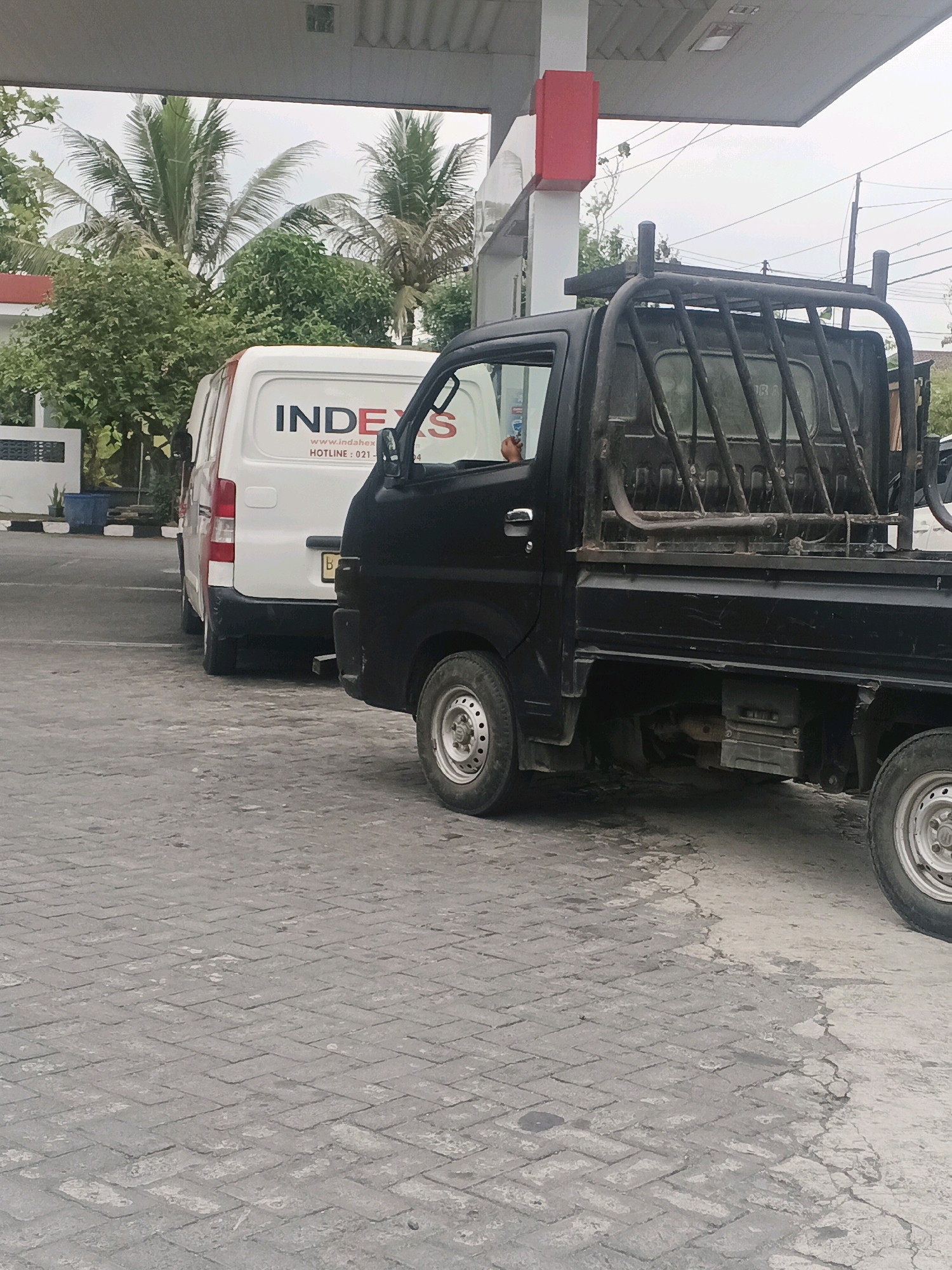 In Kliwonan, Indonesia, two vehicles are parked at a gas station. A white van with the text "INDEX S" on its side is positioned to the left, followed by a black pickup truck with a metal cage in the rear bed, to the right. The ground is paved with interlocking gray bricks, and a section of the pavement is cracked and appears wet. In the background, palm trees and lush green foliage are visible. The sky is overcast. The scene captures a moment at the gas station with the two vehicles at rest.