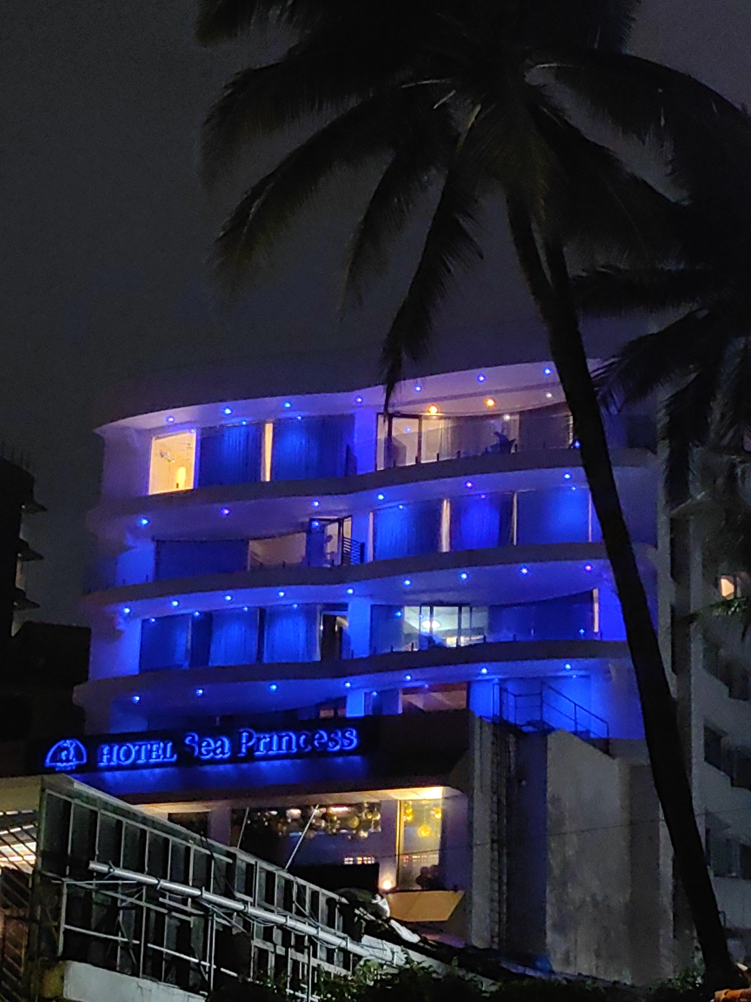 The image shows the exterior of the Hotel Sea Princess at night, illuminated by blue lights. The hotel is a multi-story building with balconies and windows. A palm tree is visible in the foreground, partially obscuring the view of the hotel. The hotel is located on the Marine Drive in Mumbai, India. The building's facade is painted white and adorned with numerous small blue lights, creating a striking contrast with the dark night sky. Some windows on the upper floors have warm, yellow light emanating from them. The hotel's sign, lit with blue neon, is clearly visible, reading "HOTEL Sea Princess." In front of the hotel, there is a metal structure, possibly a walkway or part of a fencing system, also visible in the low light. The overall scene suggests a coastal urban environment.