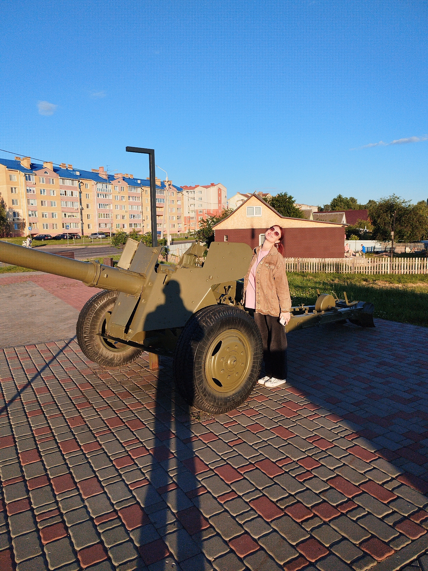 A young woman with reddish-brown hair and sunglasses stands next to a military cannon on a brick-paved area. She is wearing a light brown jacket over a pink shirt and dark pants, with white sneakers. The cannon is olive green and appears to be stationary, with large wheels. The woman is looking up and to the right, with her head tilted back. In the background, there are apartment buildings with blue roofs, a wooden fence, green grass, and trees. The sky is clear and blue, with a few wispy clouds. The scene is illuminated by the warm light of the sun, casting long shadows.